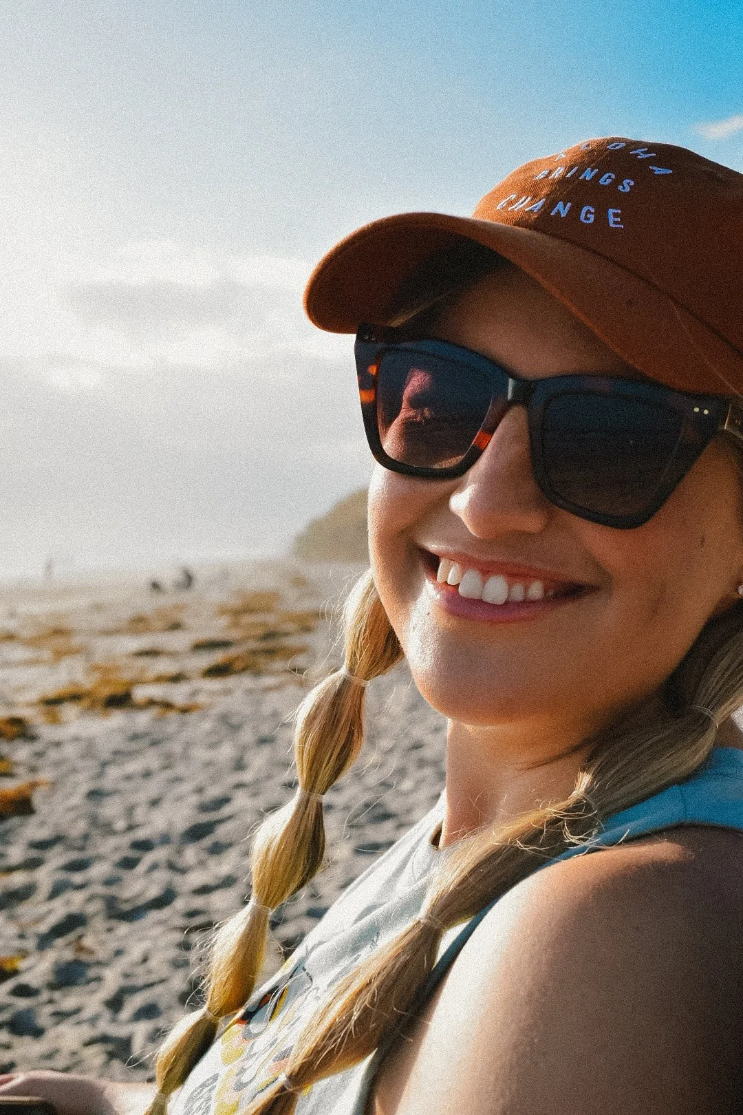 A smiling woman with braided hair wearing sunglasses and a brown cap on a beach during sunset.