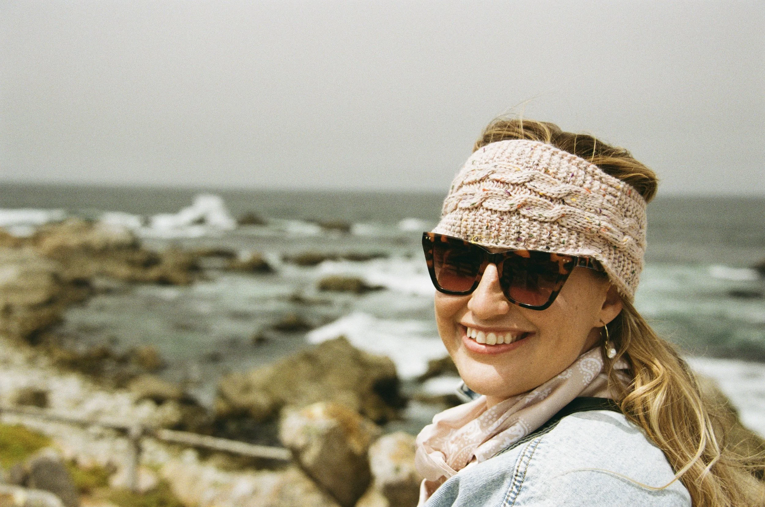 Woman wearing sunglasses, a knitted headband, and a scarf, smiling at the camera near a rocky shoreline with ocean waves.