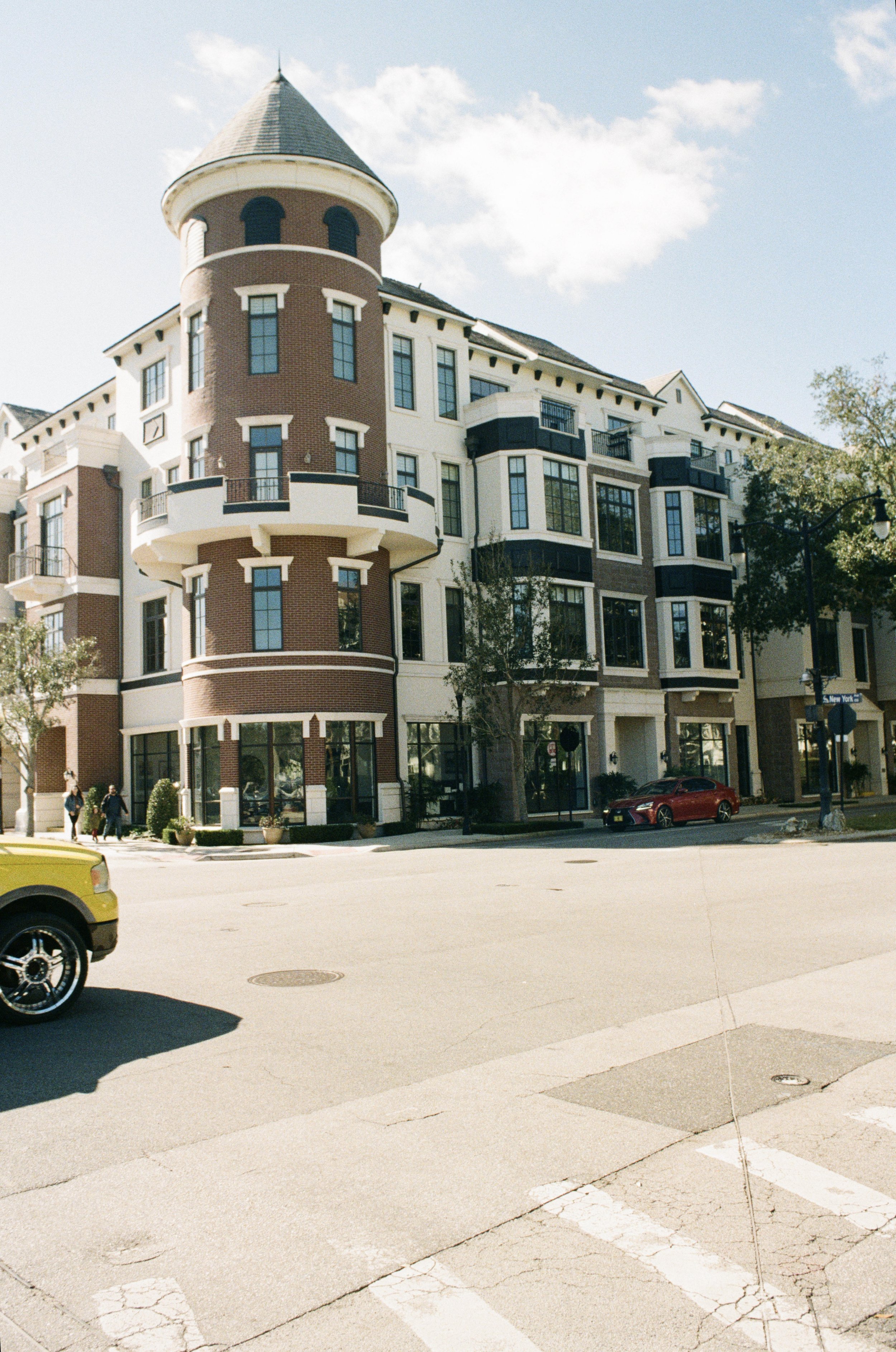 A large, multi-story residential building with a turret and balconies, located on a city street with cars and pedestrians. The sky is clear and partly cloudy.