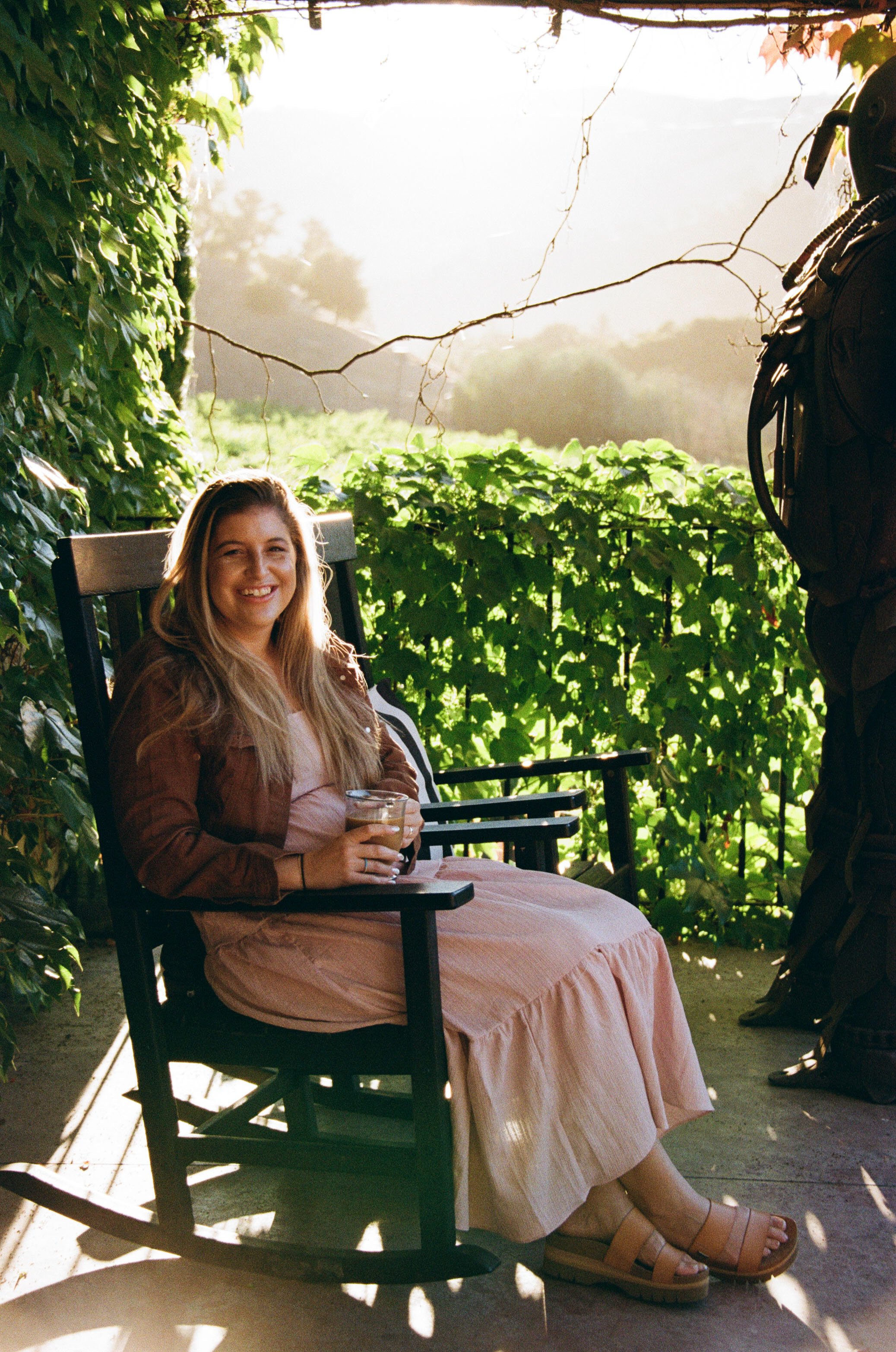 A woman sitting outdoors on a wooden chair, holding a drink, smiling, wearing a long pink dress, and tan sandals, with lush greenery and sunlight in the background.