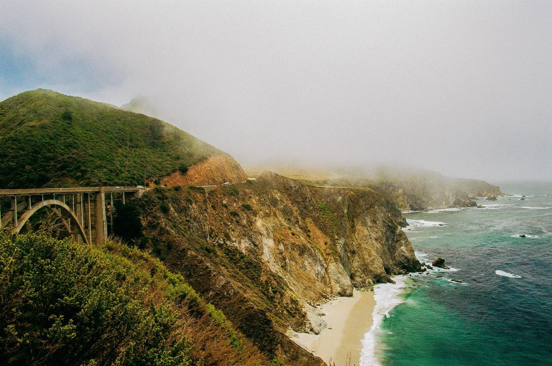 Coastal landscape showing rugged cliffs, a beach, and ocean waves, with a bridge and road running along the hillside, partially covered by fog.