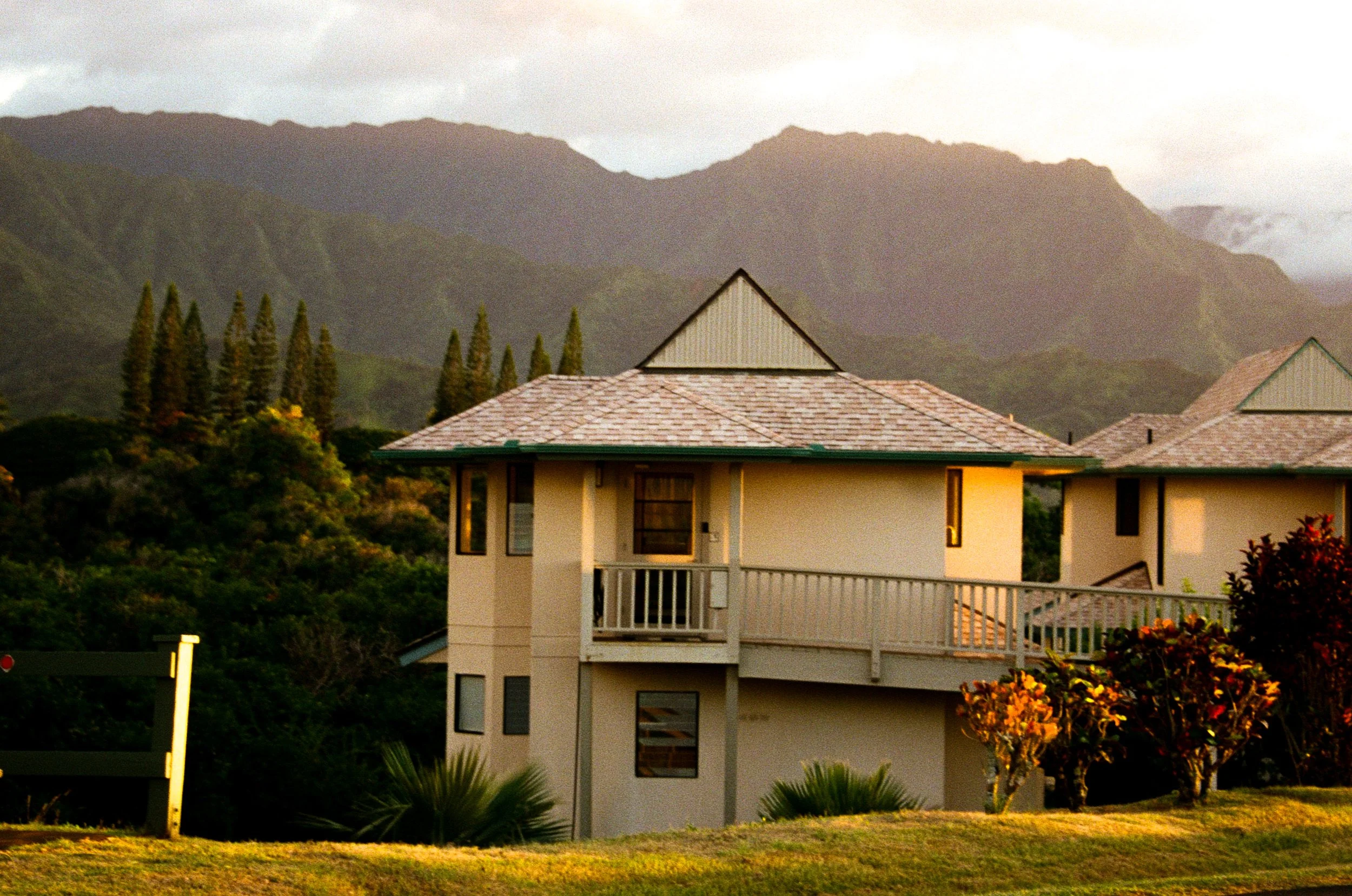 A multi-story house with beige walls and a green roof, surrounded by trees and shrubs, set against a mountainous background under cloudy skies.