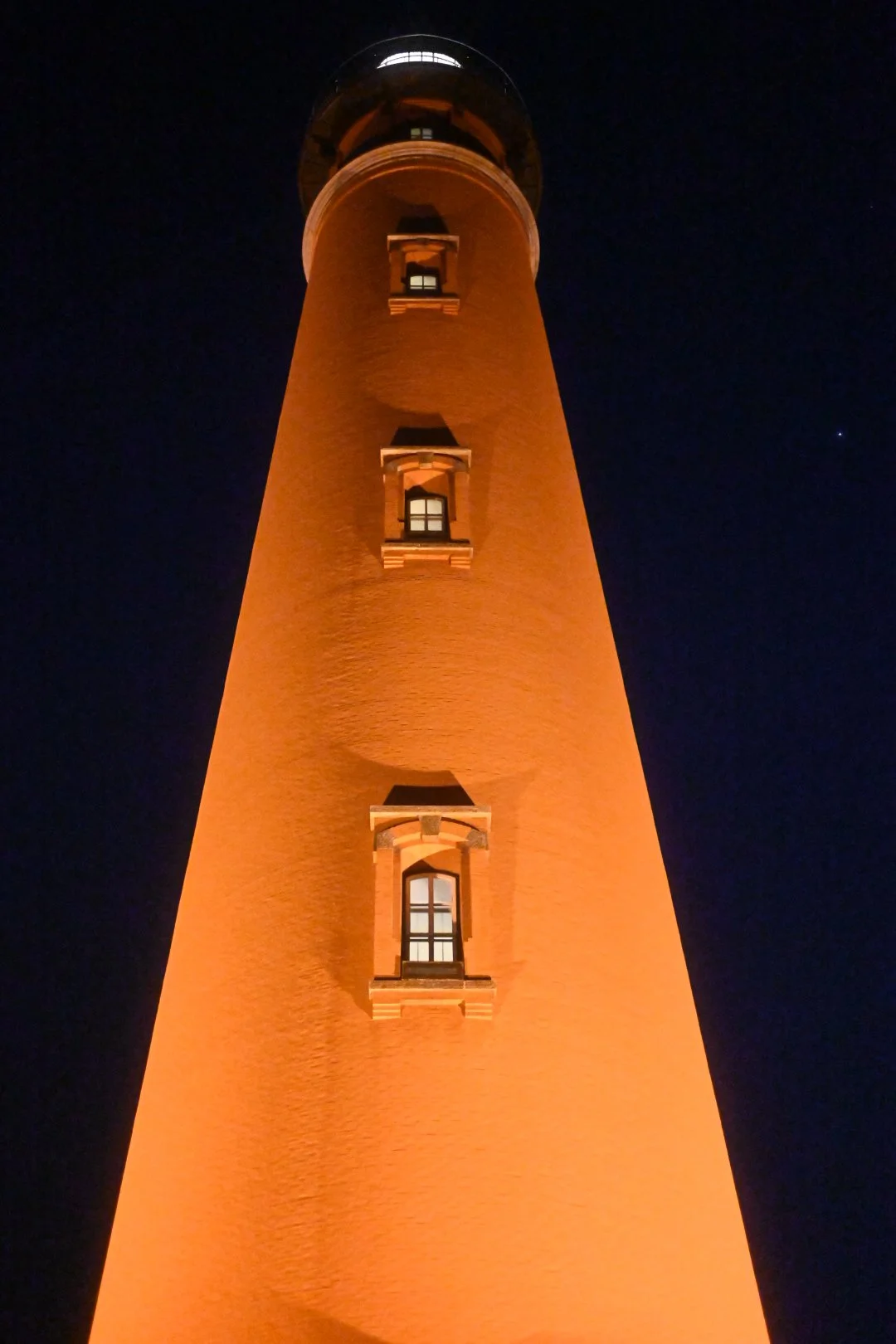 A tall lighthouse illuminated with orange lighting at night, showing three small windows and a glass observation deck at the top, set against a dark sky with a few visible stars.