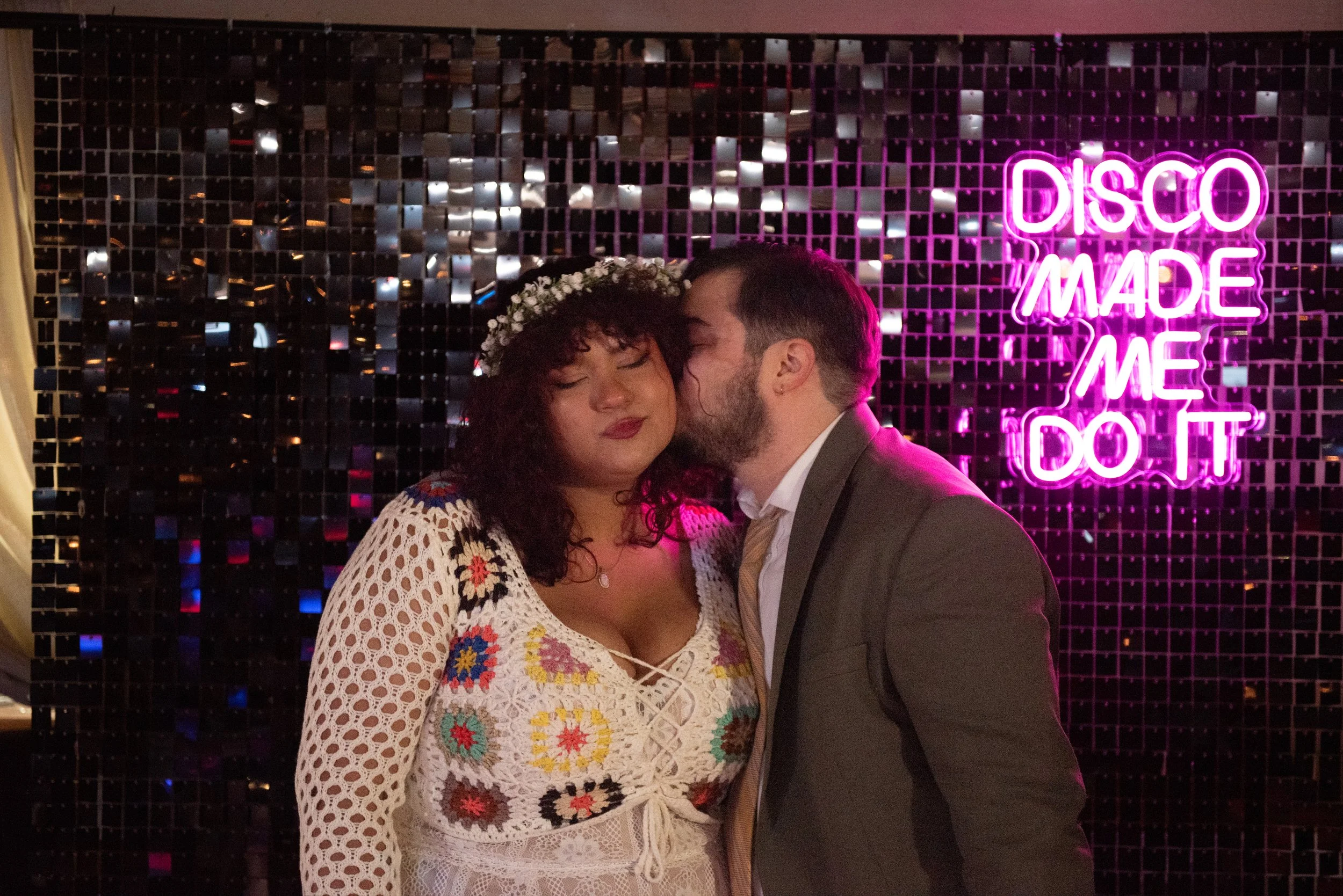 A couple stands close together in front of a dark, shiny mosaic tile wall with a neon pink sign that reads 'DISCO MADE ME DO IT'. The woman has curly hair, wears a floral crown, and a crocheted white dress with colorful flower patterns. The man has s