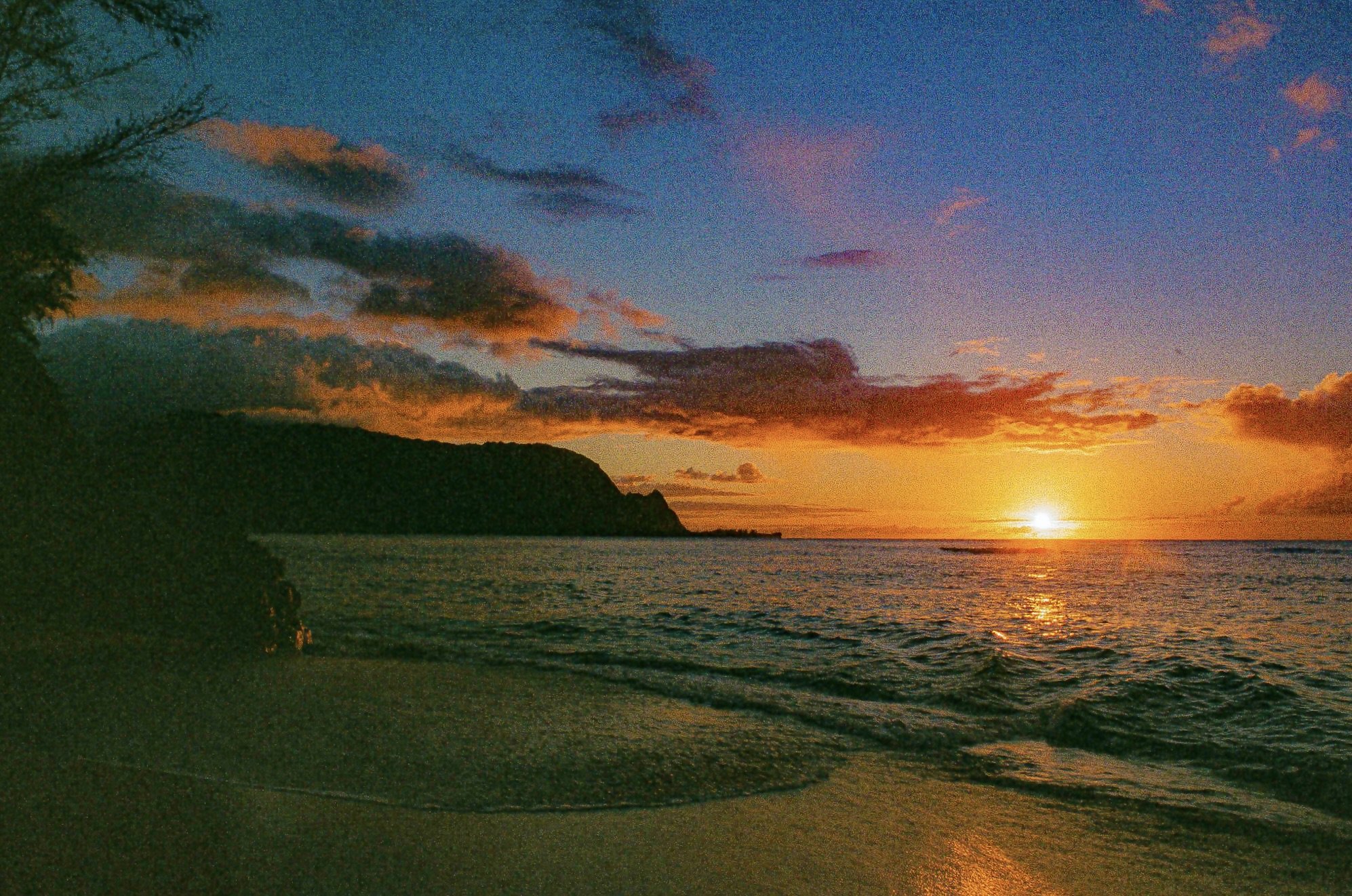 Sunset over the ocean with colorful clouds and a silhouetted coastline with trees and cliffs.