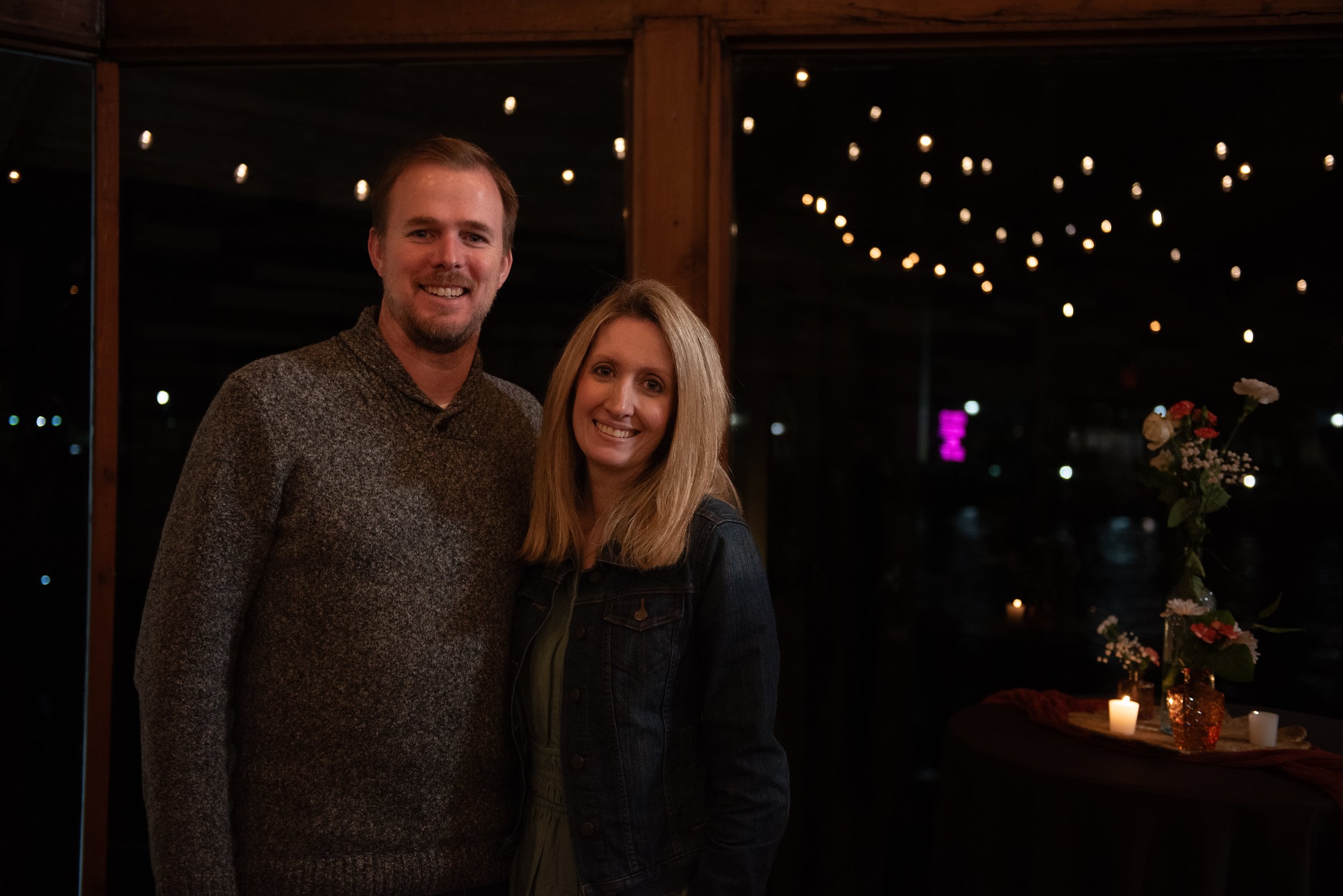 A man and woman smiling at a nighttime indoor event, background decorated with small hanging lights and flowers on a table with candles.