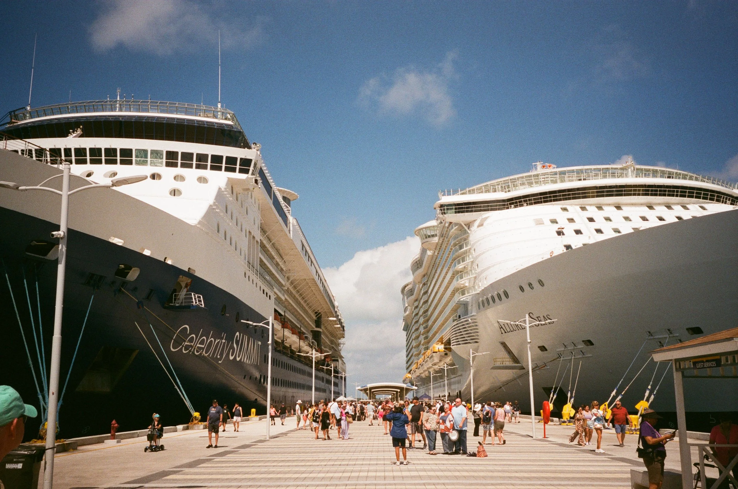 View of two large cruise ships docked at a port with a crowd of people walking on the promenade and a blue sky with some clouds overhead.
