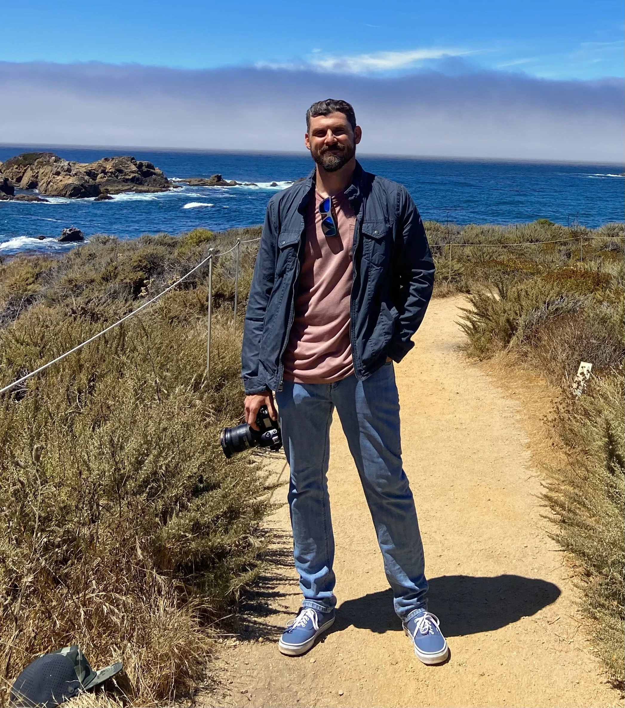 A man standing on a dirt trail near the ocean, holding a camera, wearing a gray jacket, pink t-shirt, blue jeans, and sneakers; a black helmet with a face shield is on the ground nearby. The background features rocky cliffs, blue ocean waves, and a partly cloudy sky.