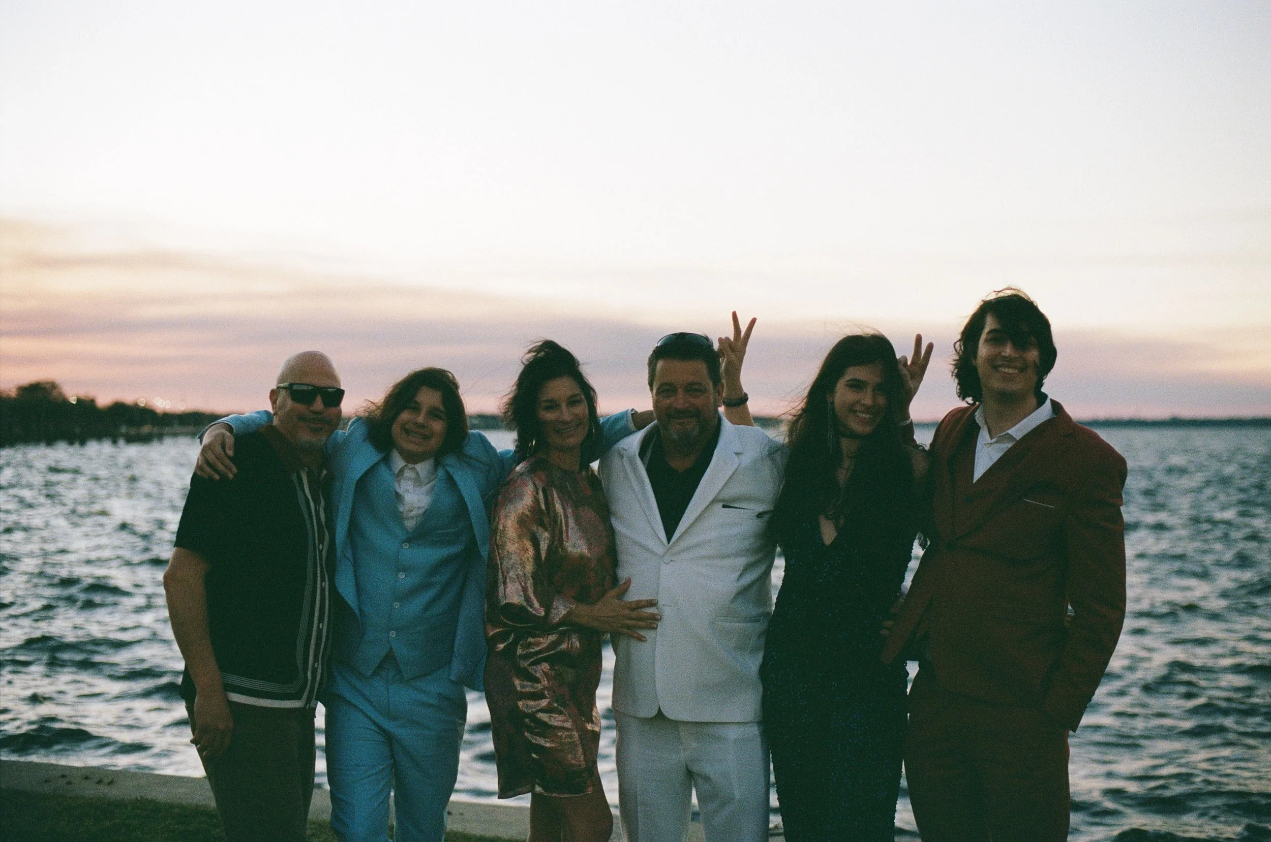 A group of six people standing together near water during sunset, smiling and making peace signs.