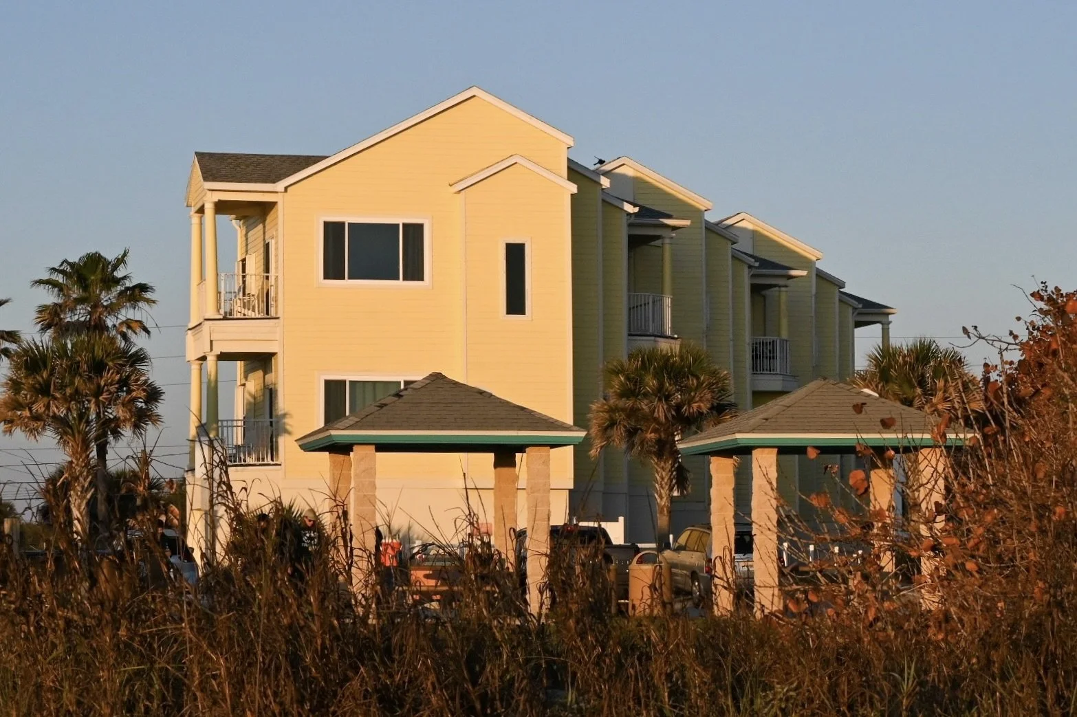 Multi-story yellow residential building with balconies, surrounded by palm trees and a parking area with cars, during late afternoon.