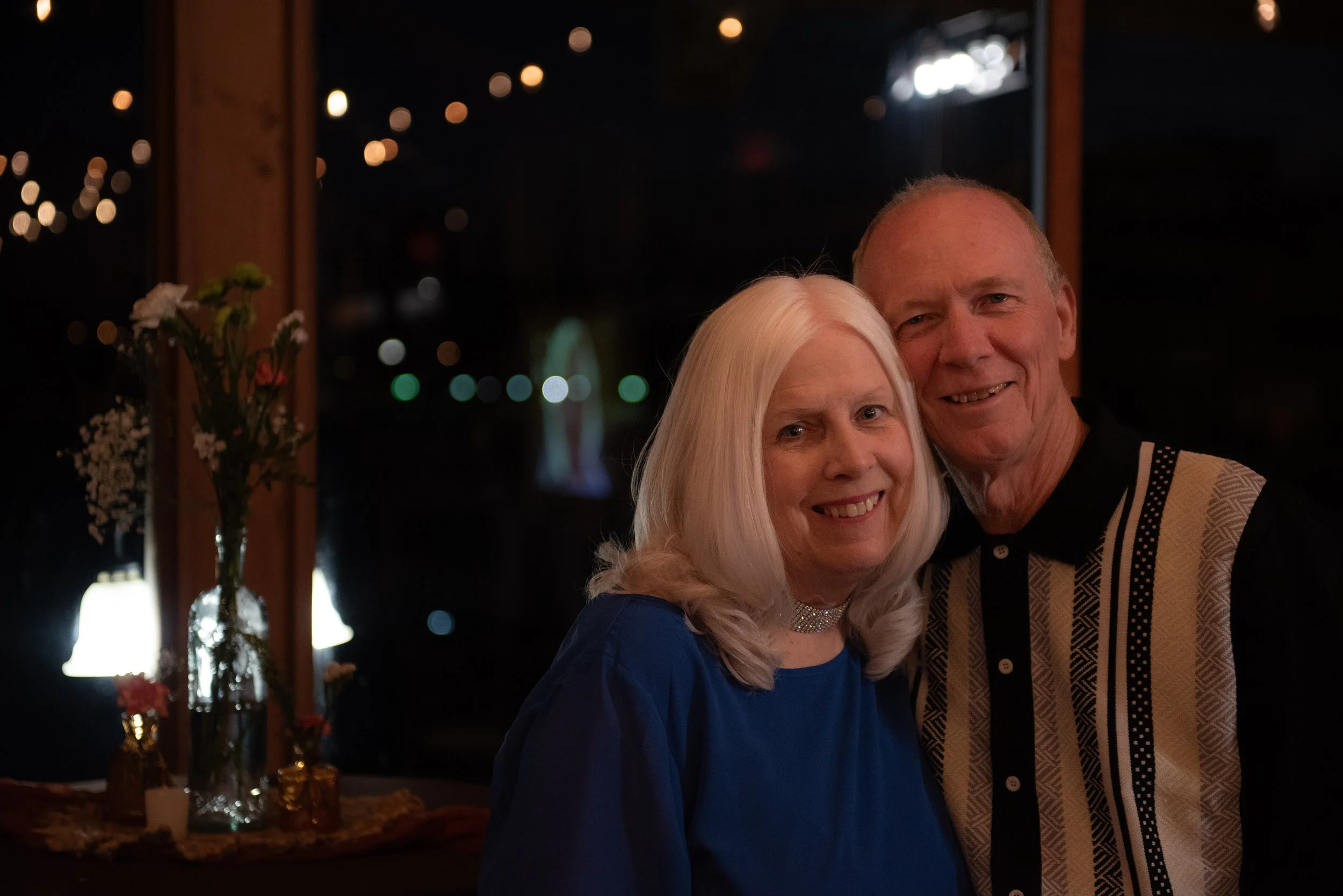 An elderly couple smiling and posing for a photo at a dimly lit indoor event with floral decorations and blurred lights in the background.