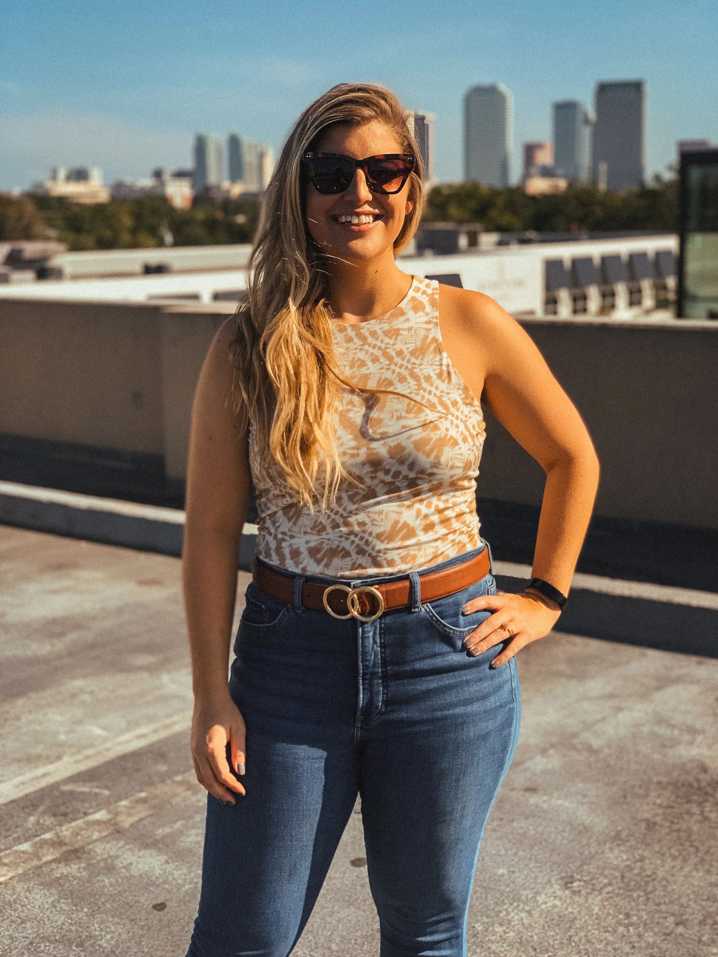 A woman with long blonde hair wearing sunglasses, a sleeveless animal print top, a brown belt, and jeans, standing in a parking lot with a city skyline in the background on a sunny day.