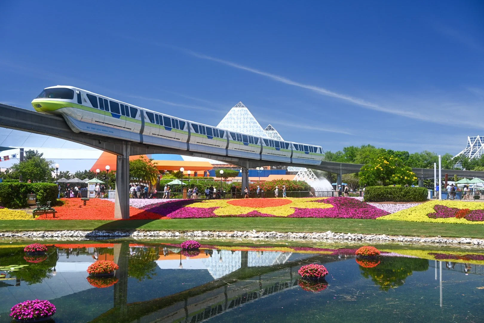 A Disney theme park with a reflective water feature, colorful flower beds, and a monorail passing overhead under a bright blue sky with a few wispy clouds.