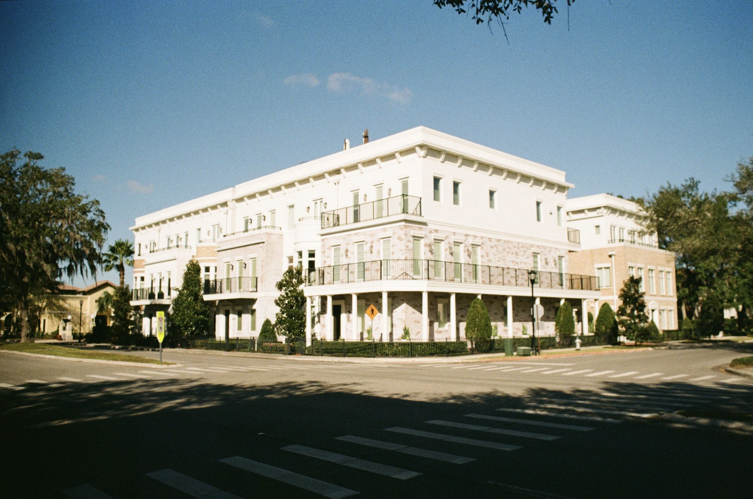 A white multi-story building with balconies and large windows is situated on a street corner, surrounded by trees and a crosswalk visible on the street in front.