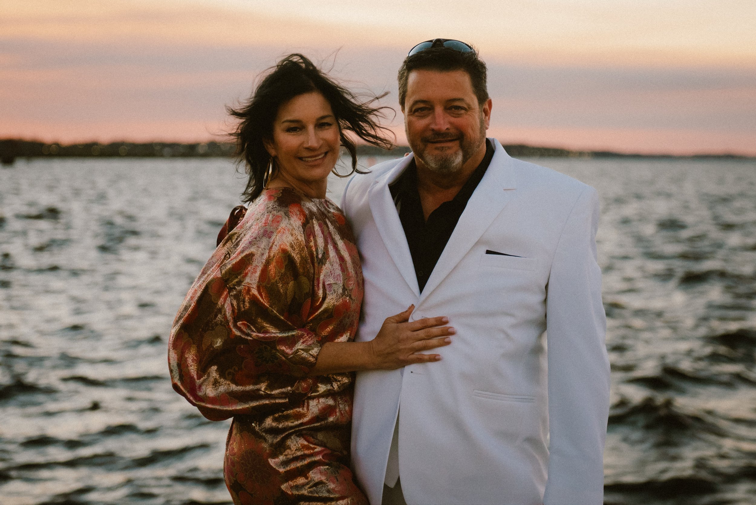 A smiling couple standing close together near a body of water at sunset, with the woman in a colorful patterned dress and the man in a white suit jacket.
