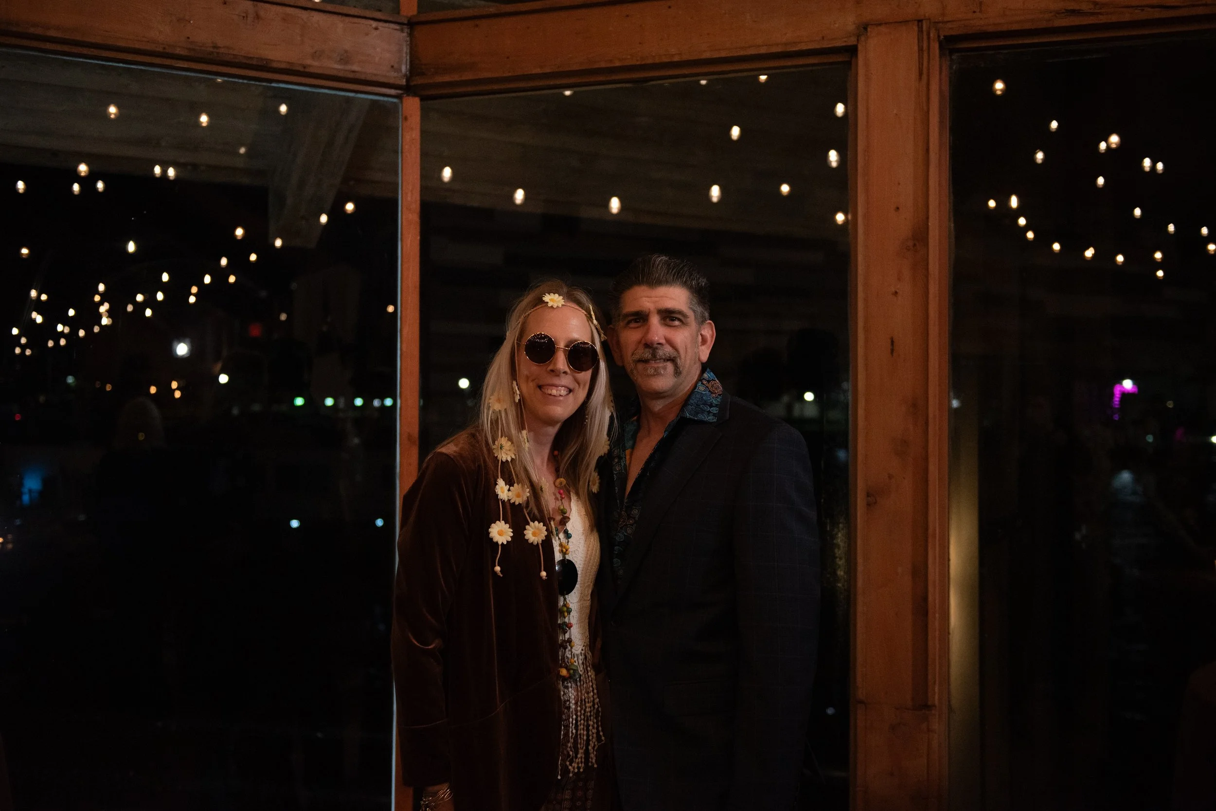 A man and woman are standing together inside at night, in front of a large mirror with indoor string lights reflected in the glass, visible behind them.