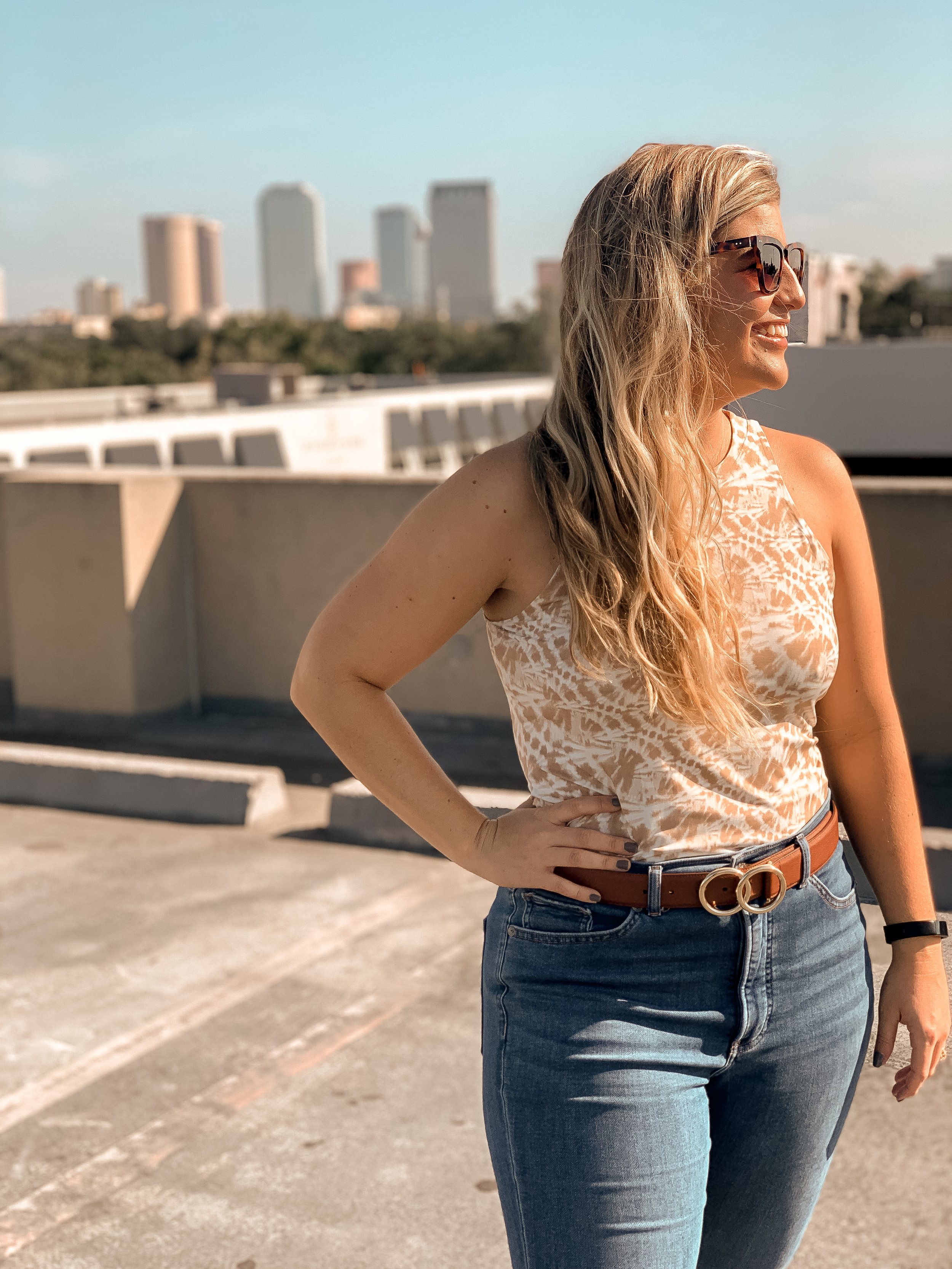 Side view of a smiling woman wearing glasses, a sleeveless animal print top, a brown belt, and jeans, standing on a rooftop parking lot with city skyscrapers in the background.