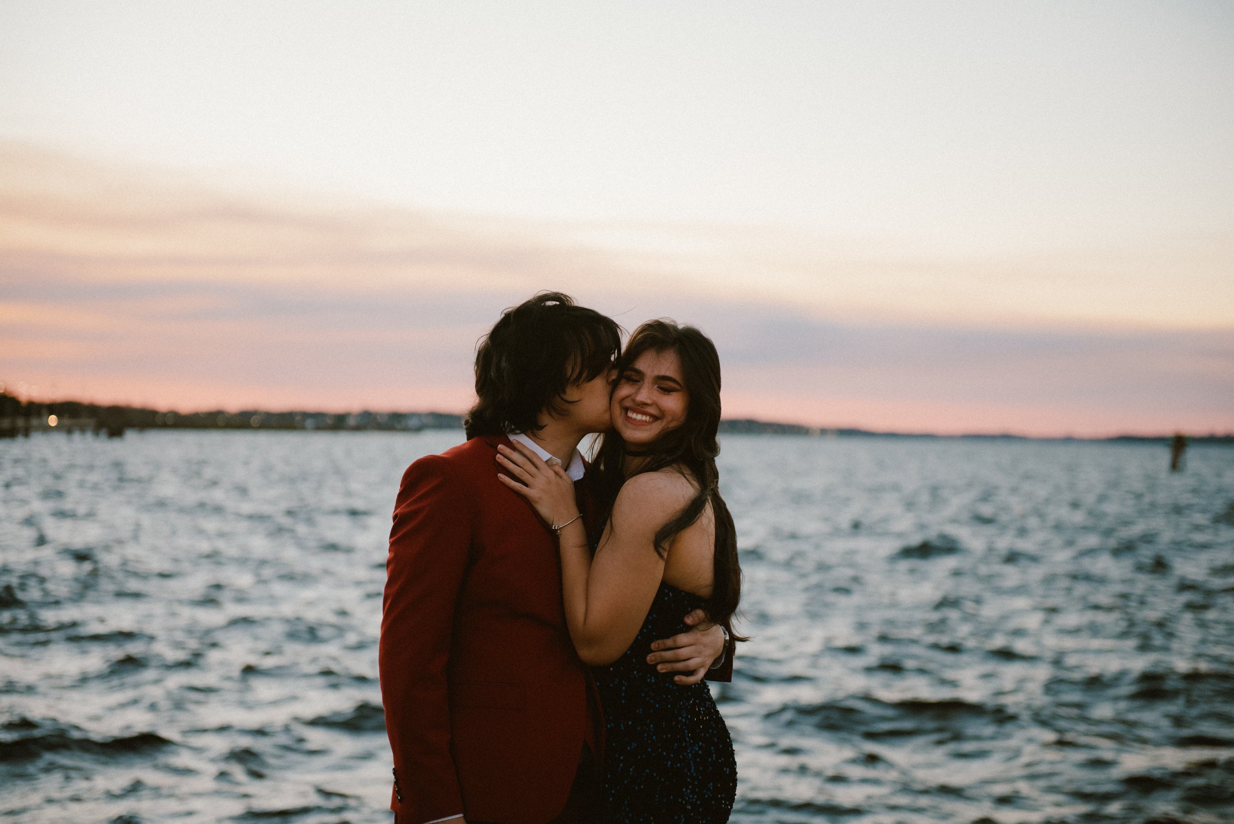 A young couple is embracing and smiling by a body of water during sunset. The man is kissing the woman's cheek, and the woman has her eyes closed with a joyful expression.