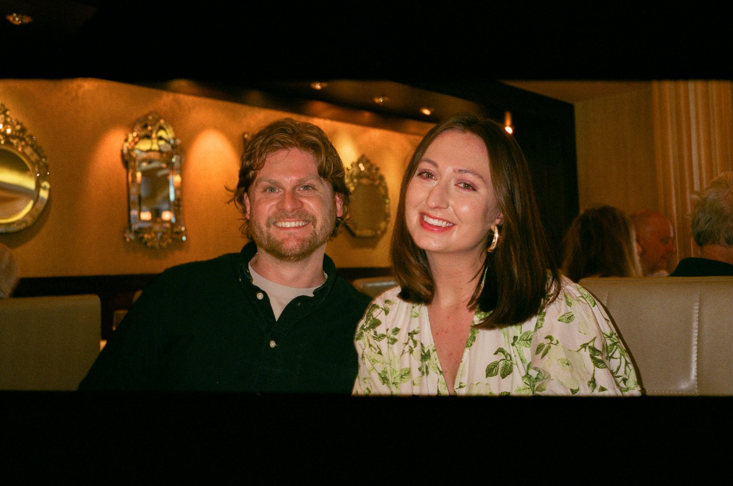 A man with brown, wavy hair and a beard, wearing a black shirt, and a woman with long, straight dark hair, wearing a white dress with green leaf patterns, smiling and sitting in a warmly lit restaurant with ornate gold-framed mirrors on the wall behi