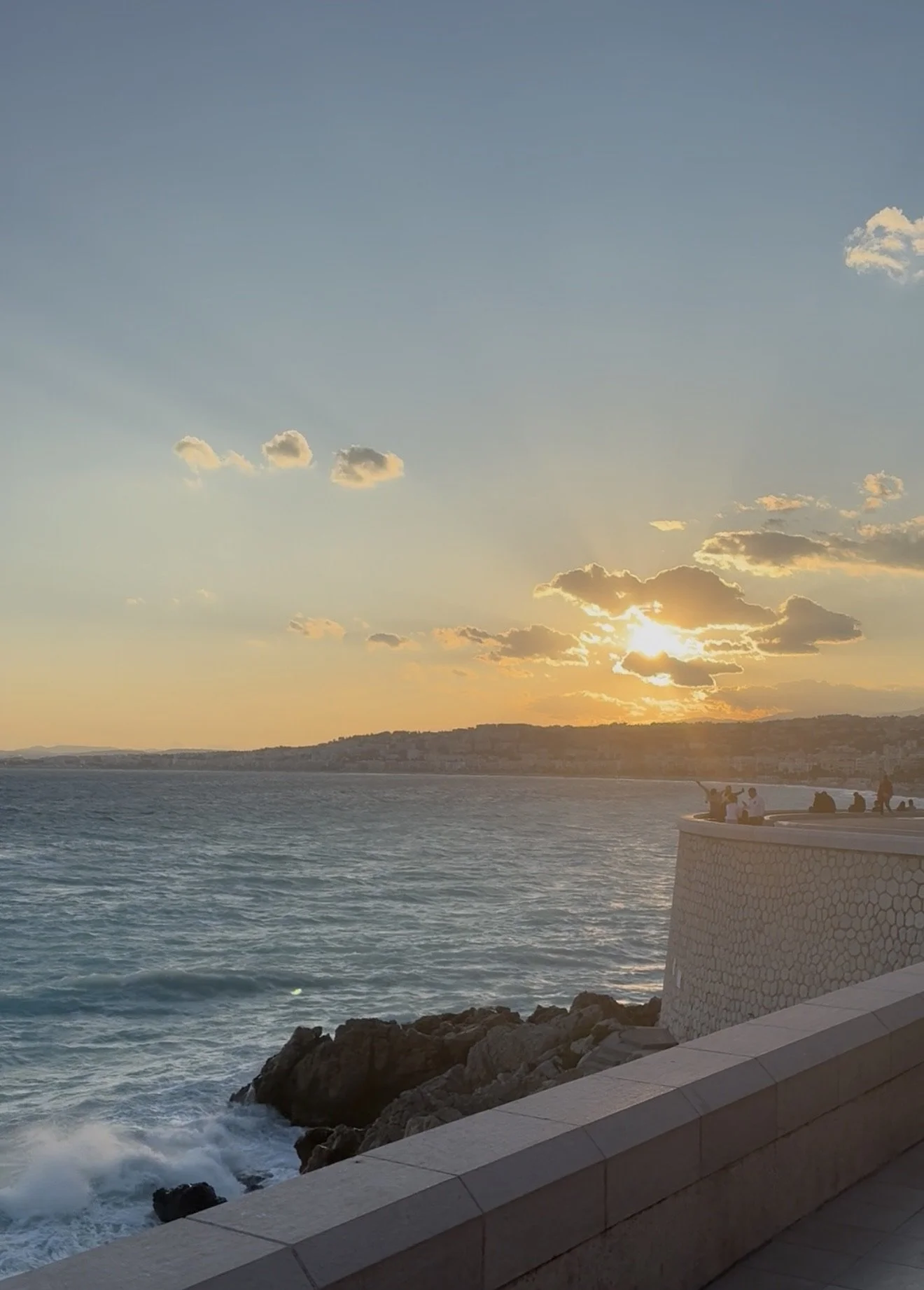 Sunset over the ocean with a promenade and people sitting on the railing, and a mostly clear sky with a few scattered clouds.
