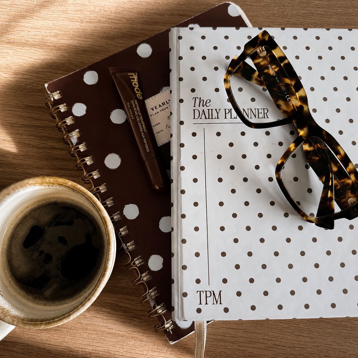 Glass of black coffee, hardcover polka dot planner, smaller polka dot notebook, tortoise shell sunglasses, and a lipstick tube on a wooden surface.