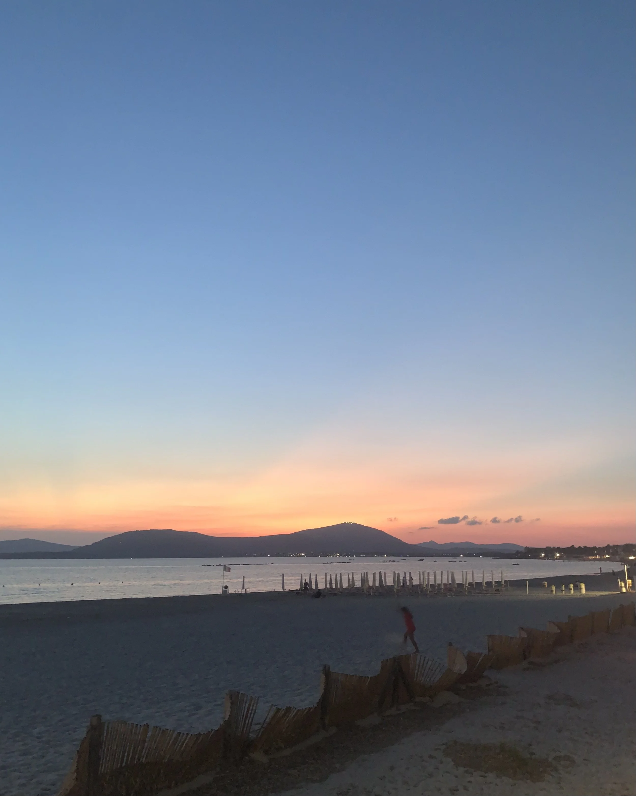 Beach scene at dusk with calm water, mountains in the background, a sky with fading sunset colors, and a few people walking near the shoreline. Beach umbrellas and chairs are present along the shore.