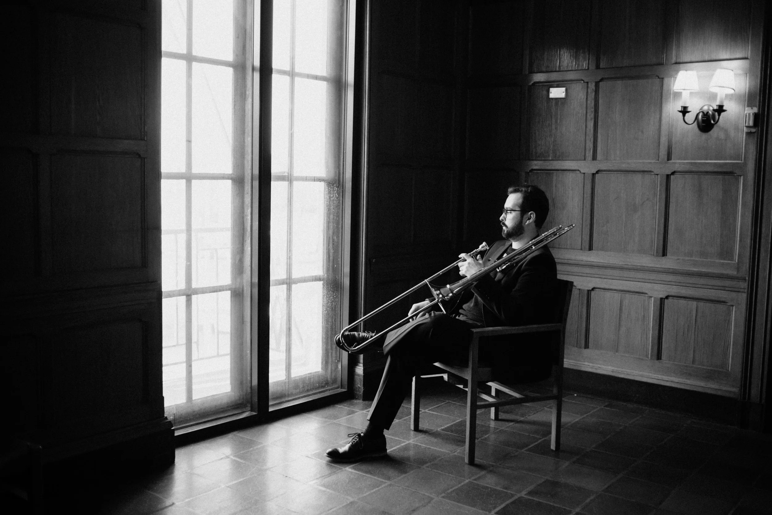 A man sitting on a chair in a music room, holding a trombone, with large windows and wood-paneled walls, in black and white.