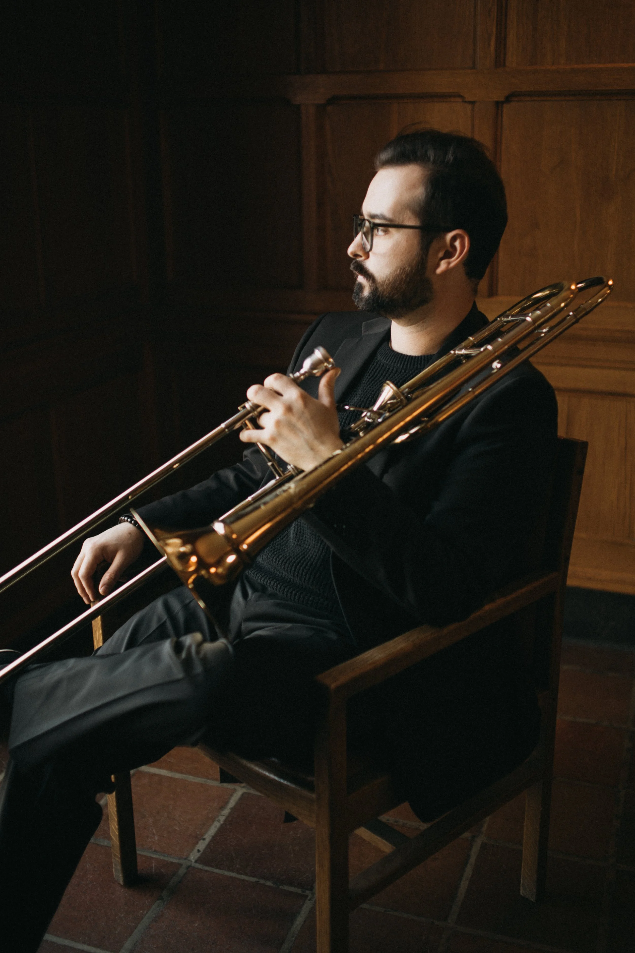 A man with glasses and a beard is sitting in a wooden chair, holding a trombone, with a wooden-paneled wall in the background.