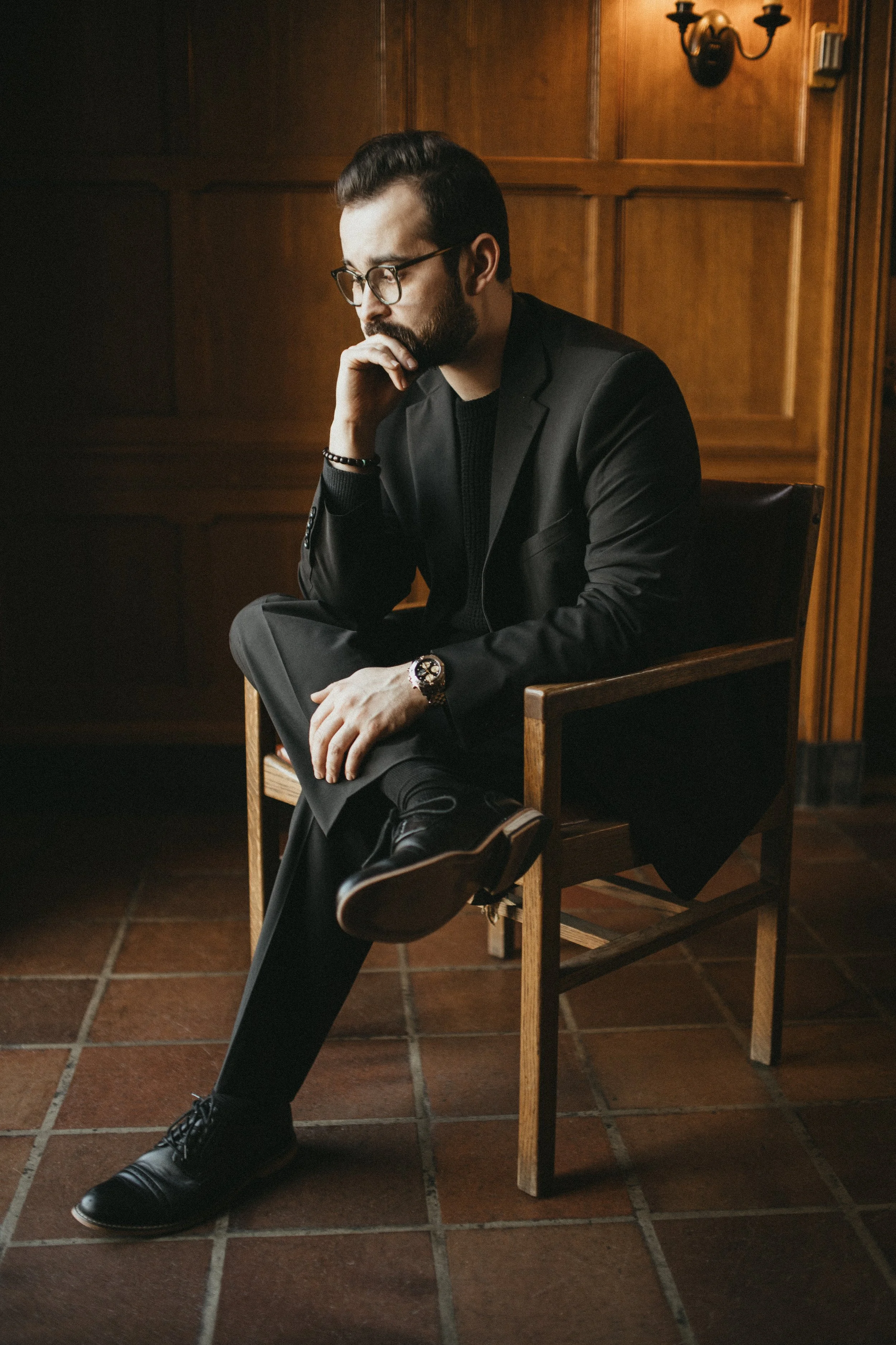 A man with dark hair, glasses, and a beard sitting in a wooden chair with one hand on his chin, wearing a black blazer, black sweater, black pants, and black shoes in a room with wood-paneled walls and tiled flooring.