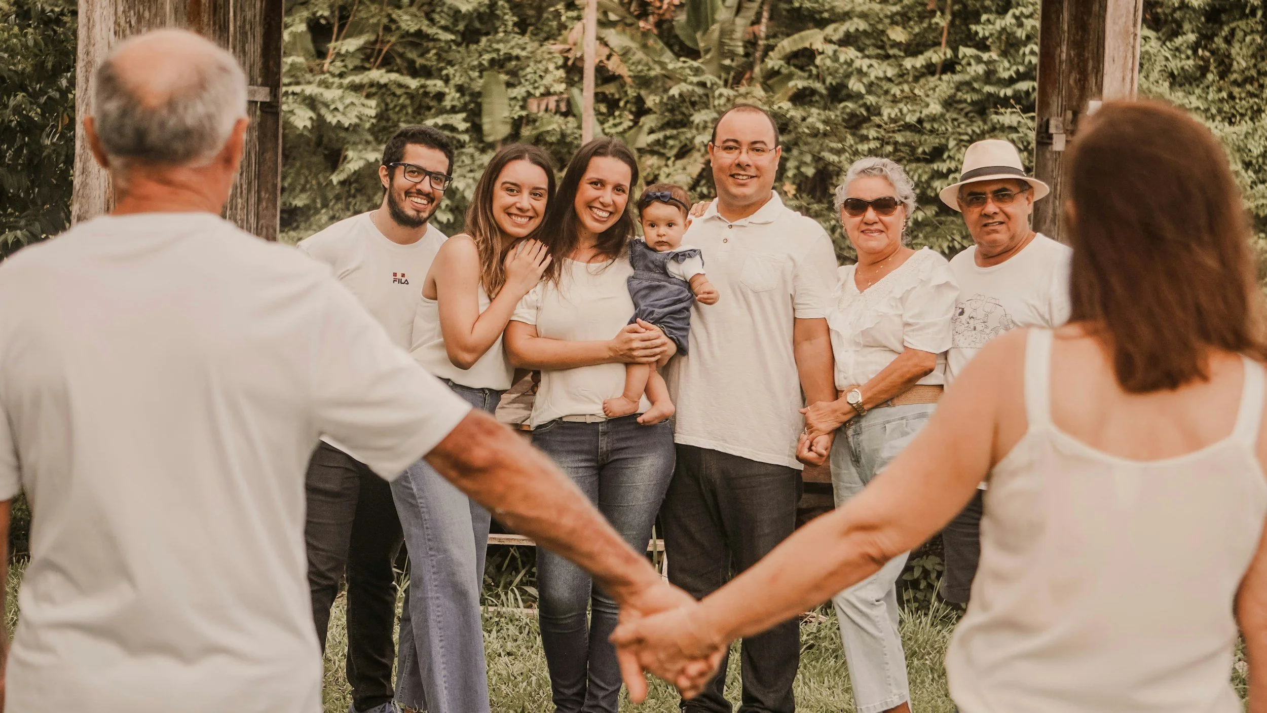Un groupe de huit personnes, adultes et un bébé, debout en cercle dans un environnement naturel. Le groupe semble heureux et engagé dans une activité sociale ou familiale, se tenant par les mains ou en interaction. L'arrière-plan est composé d'arbres et de végétation verdoyante.
