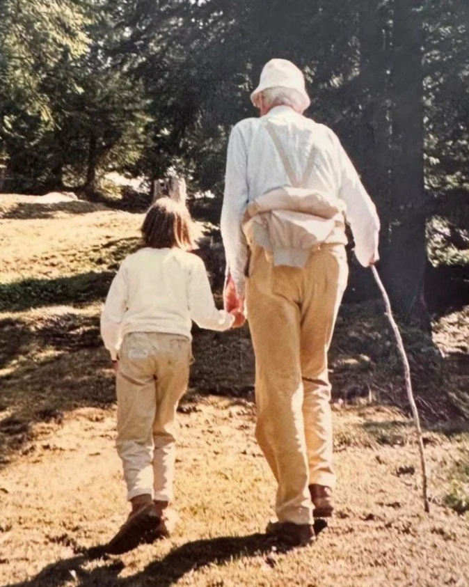 Deux personnes, une adulte et un enfant, marchant main dans la main dans un environnement herbeux avec des arbres en arrière-plan. L'adulte porte un chapeau blanc, une chemise blanche, un pantalon beige, et tient un bâton.