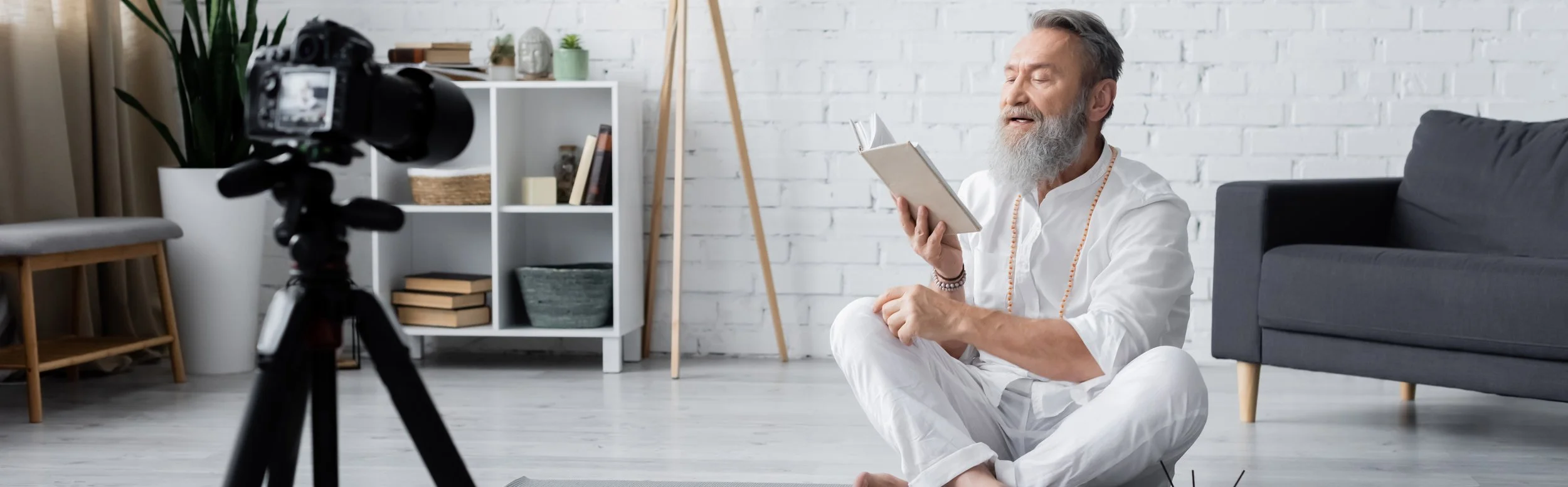 Un homme âgé avec une barbe grise, habillé en blanc, assis au sol dans un salon moderne, lisant un livre.
