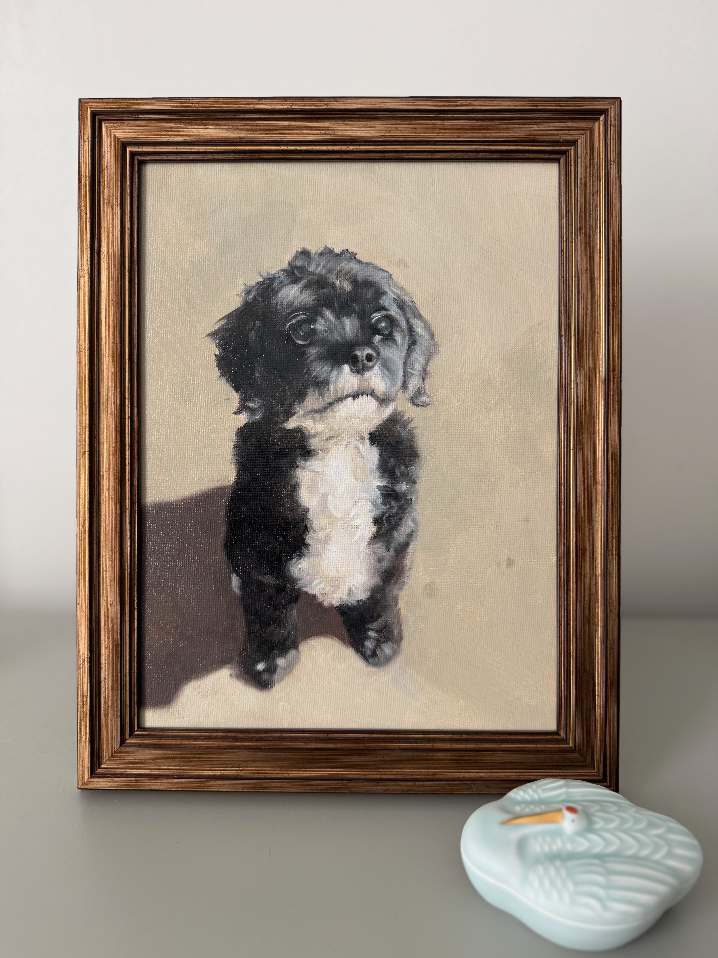 A framed painting of a black and white Cavapoo sitting against a neutral background, hanging on a white wall, with a decorative ceramic dish shaped like a swan in front of it.