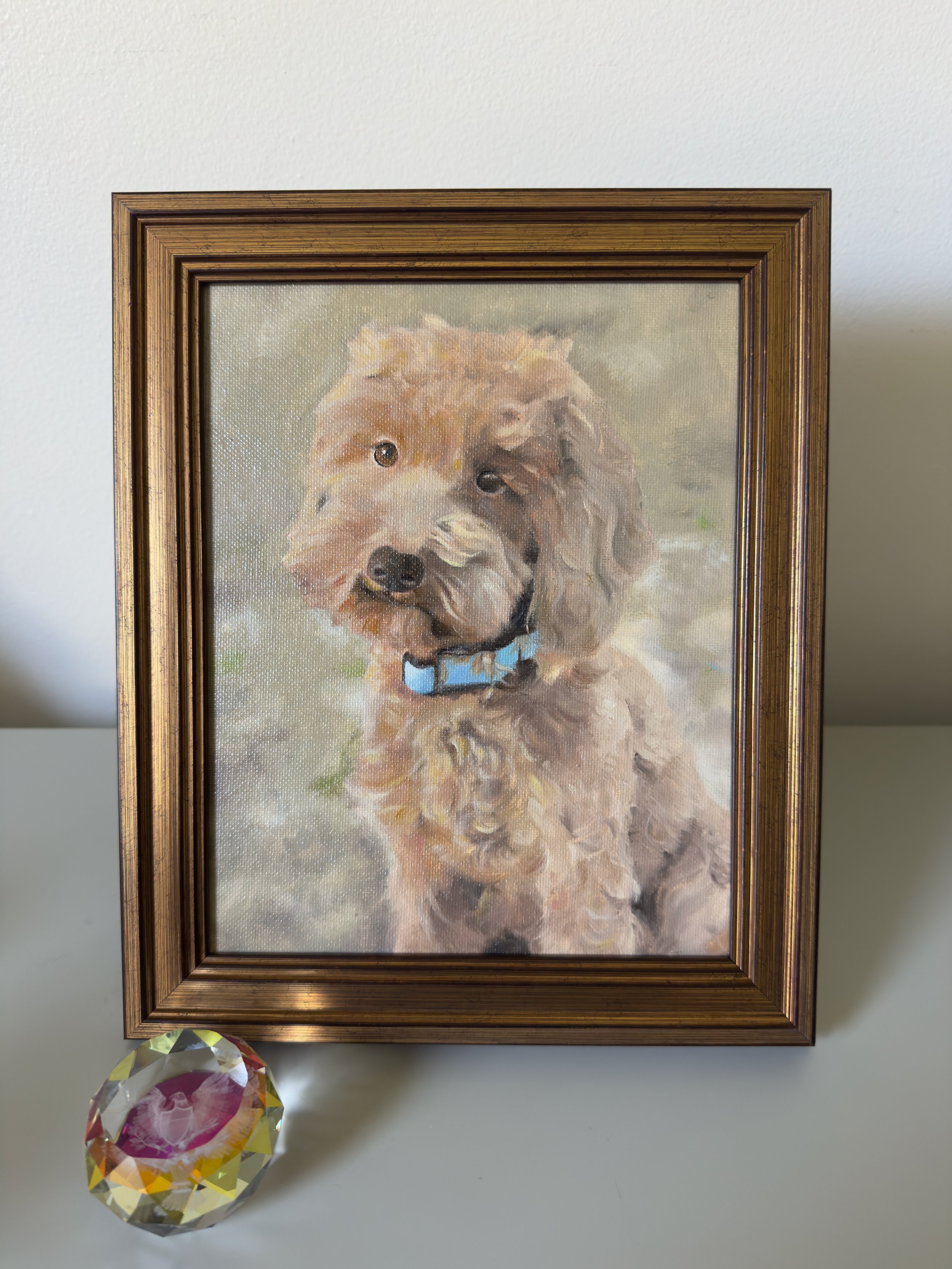 A framed watercolor painting of a dog with curly fur wearing a blue collar, hanging on a white wall. Below the painting, on a gray surface, there is a multicolored faceted crystal ball.