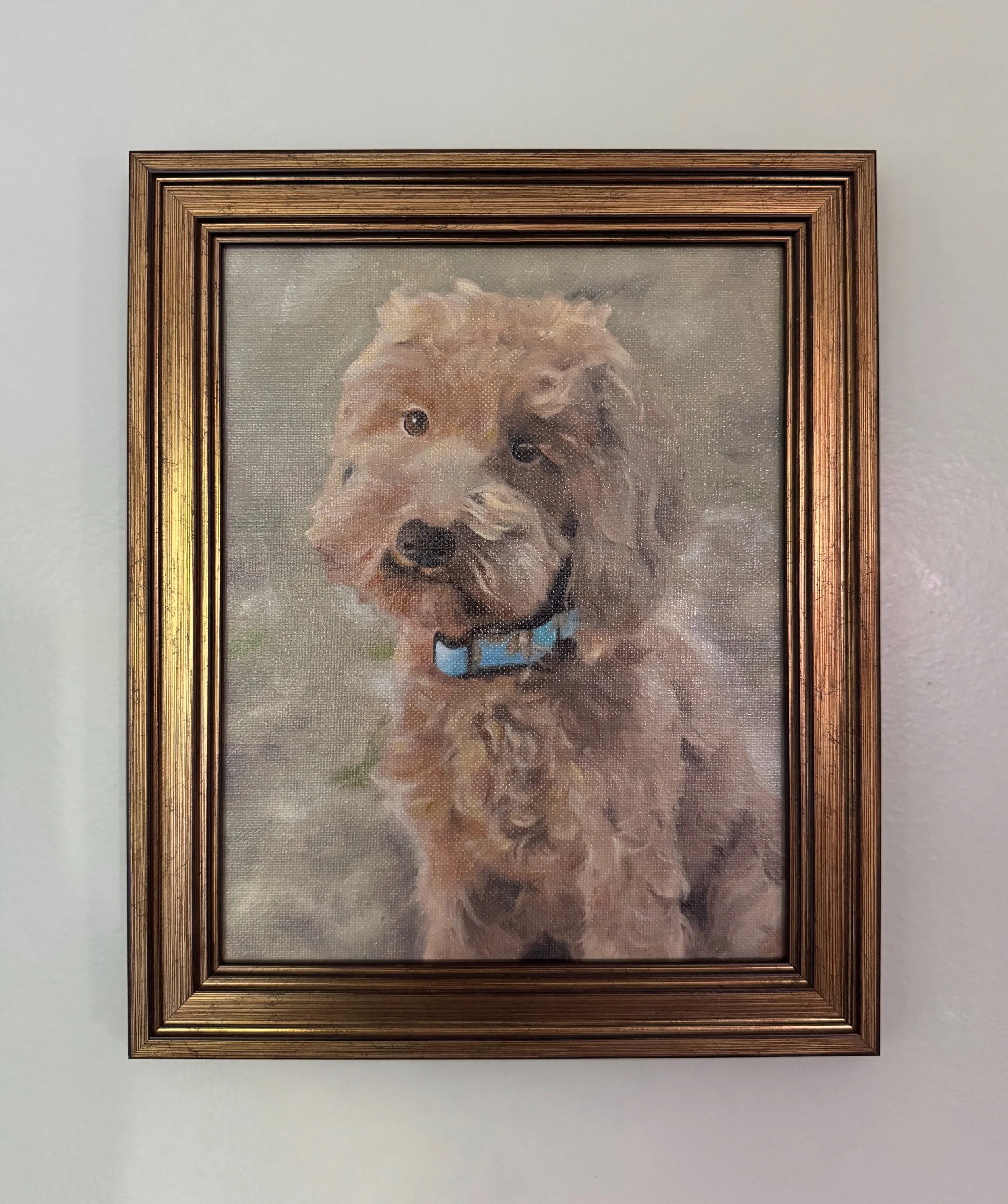 Painting of a mini Labradoodle with a curly coat, wearing a blue and black striped bow tie, within a gold-colored frame, hanging on a white wall.