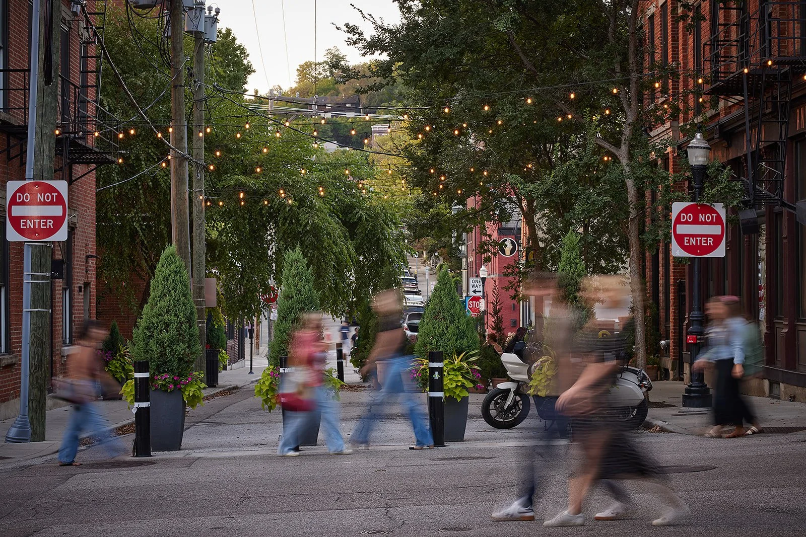 People walking on a city street, several appearing blurred due to motion, lined with trees, potted plants, brick buildings, and hanging string lights, with a motorcycle parked along the sidewalk, and several 'Do Not Enter' signs visible.