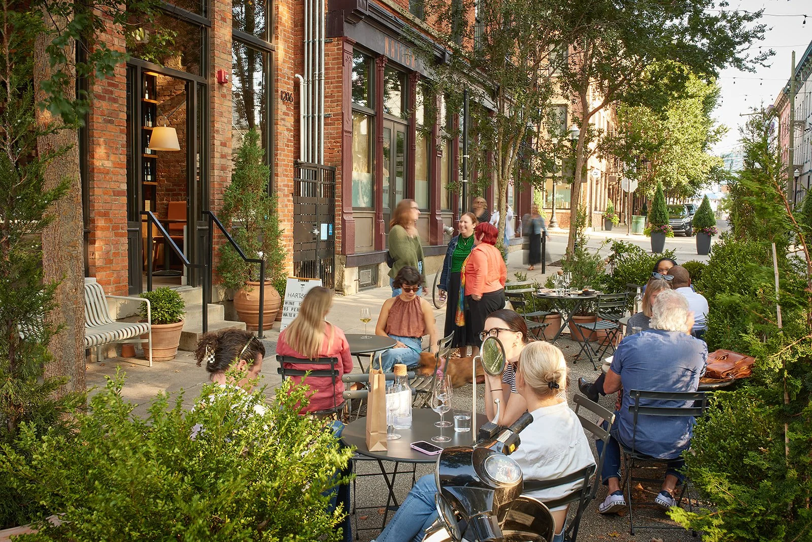 Outside patio of a restaurant or cafe with people sitting at tables, some chatting and enjoying drinks, and others standing and talking, surrounded by greenery and brick buildings.