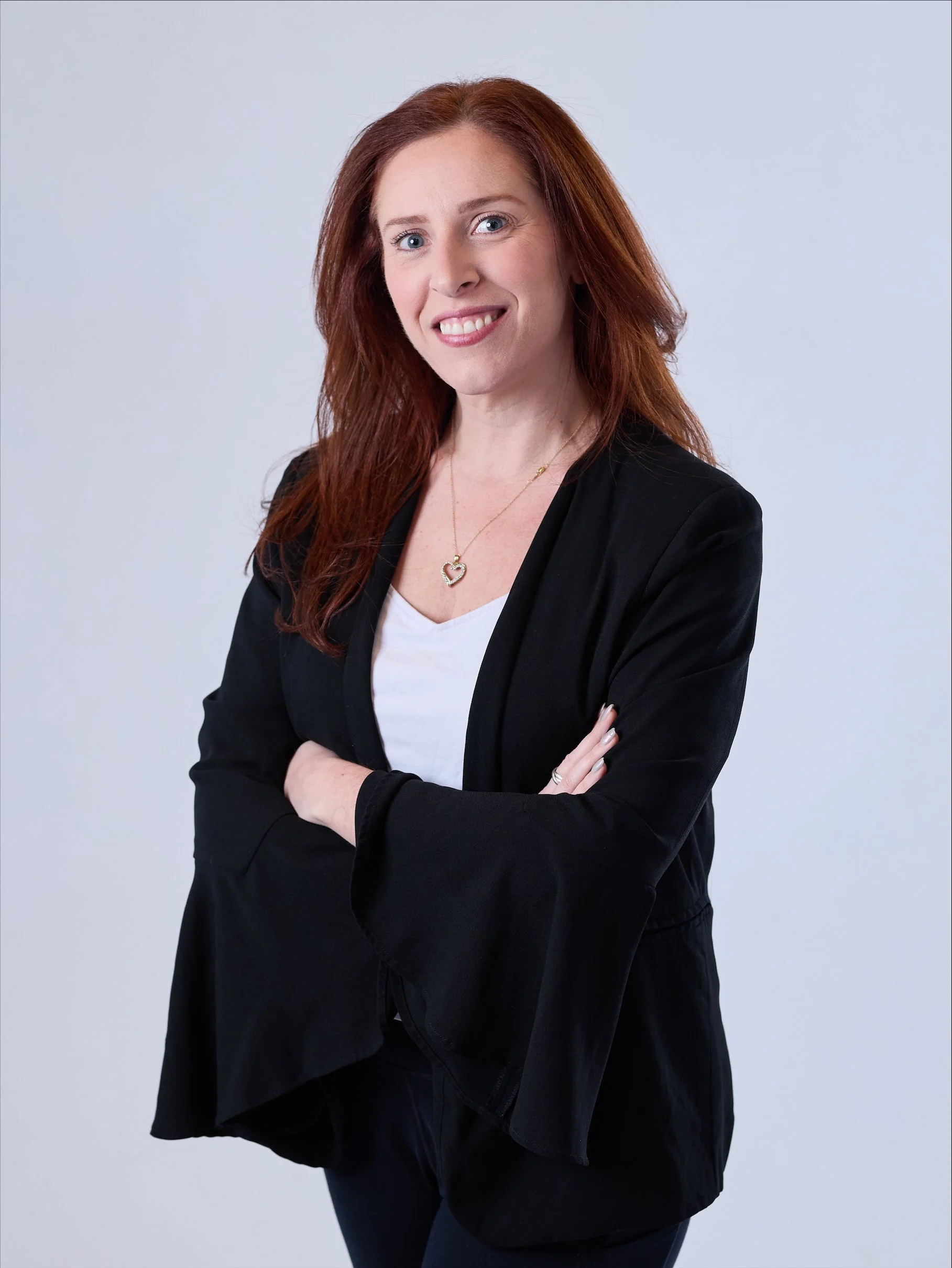 A woman with red hair, wearing a black blazer, white top, and a heart-shaped necklace, smiling with arms crossed against a plain gray background.