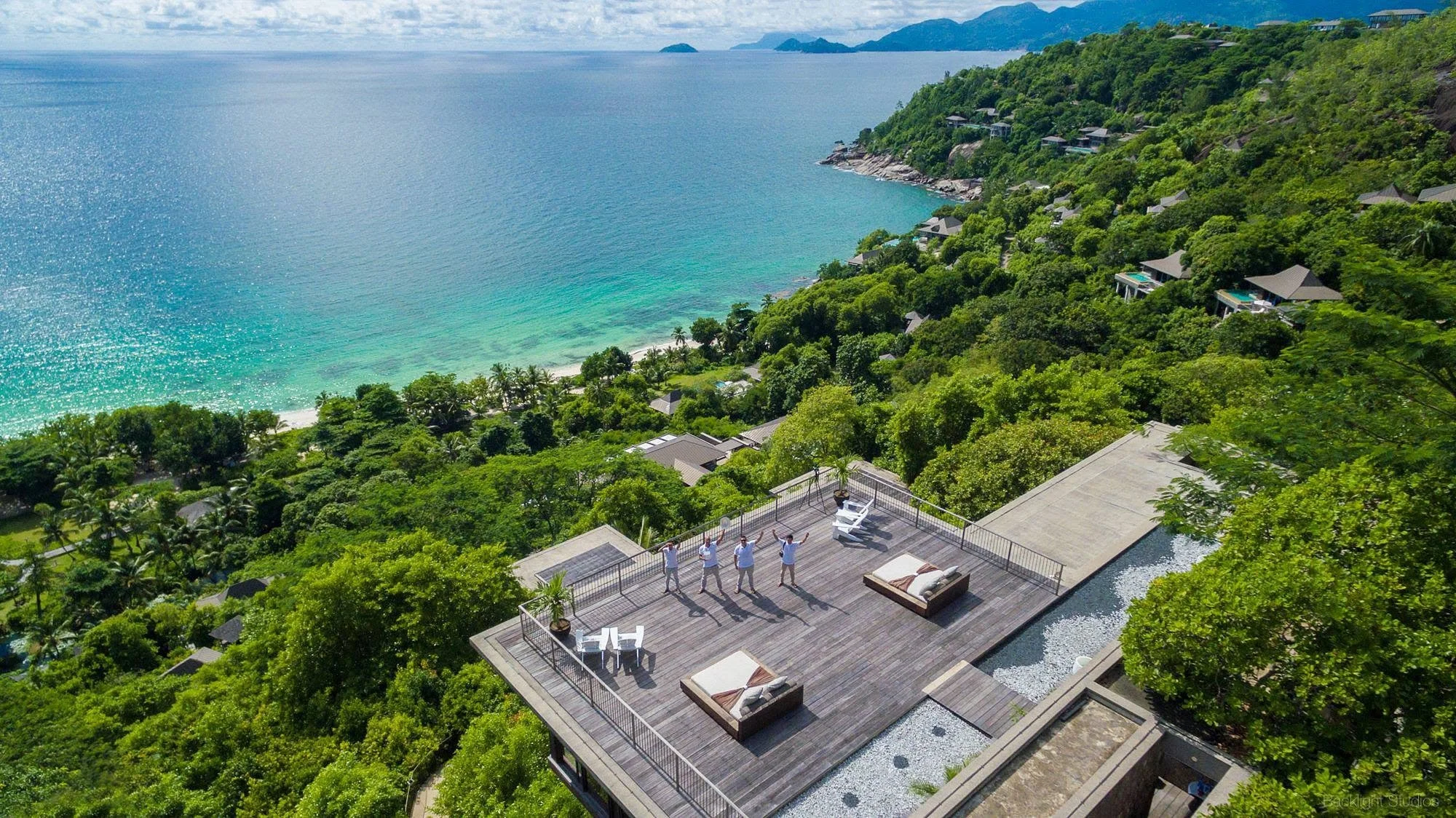 Aerial view of a large wooden deck with lounge areas and umbrellas overlooking turquoise ocean and lush green hillside with houses, mountains in the distance, and four people standing on the deck with arms raised.