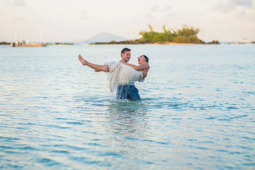 A happy couple in wedding attire enjoying a moment in the ocean, with the man holding the woman in his arms and both smiling.