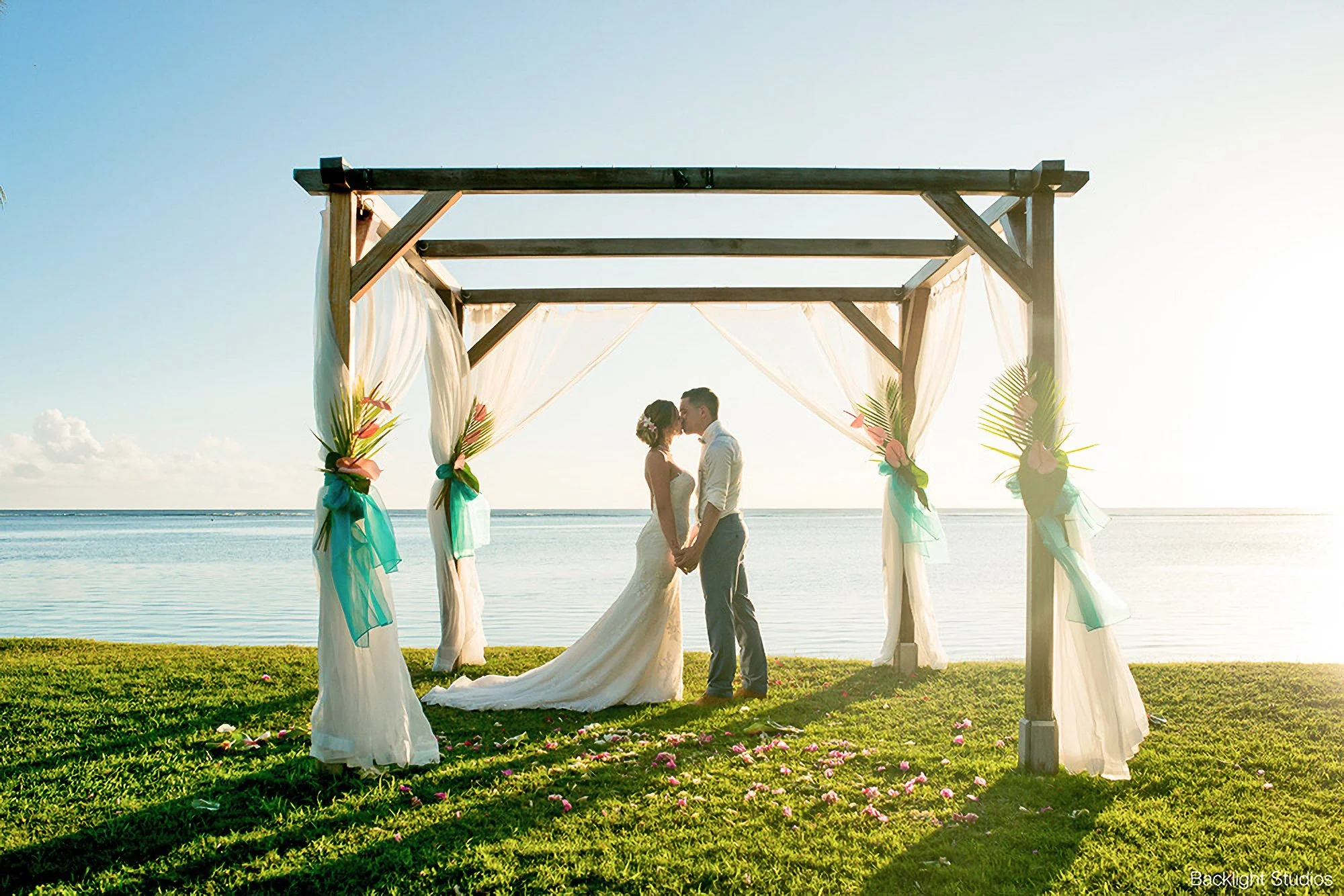 A bride and groom standing and kissing under a decorated wooden arch at a beach wedding, with water and sky in the background, and flower petals on the ground.