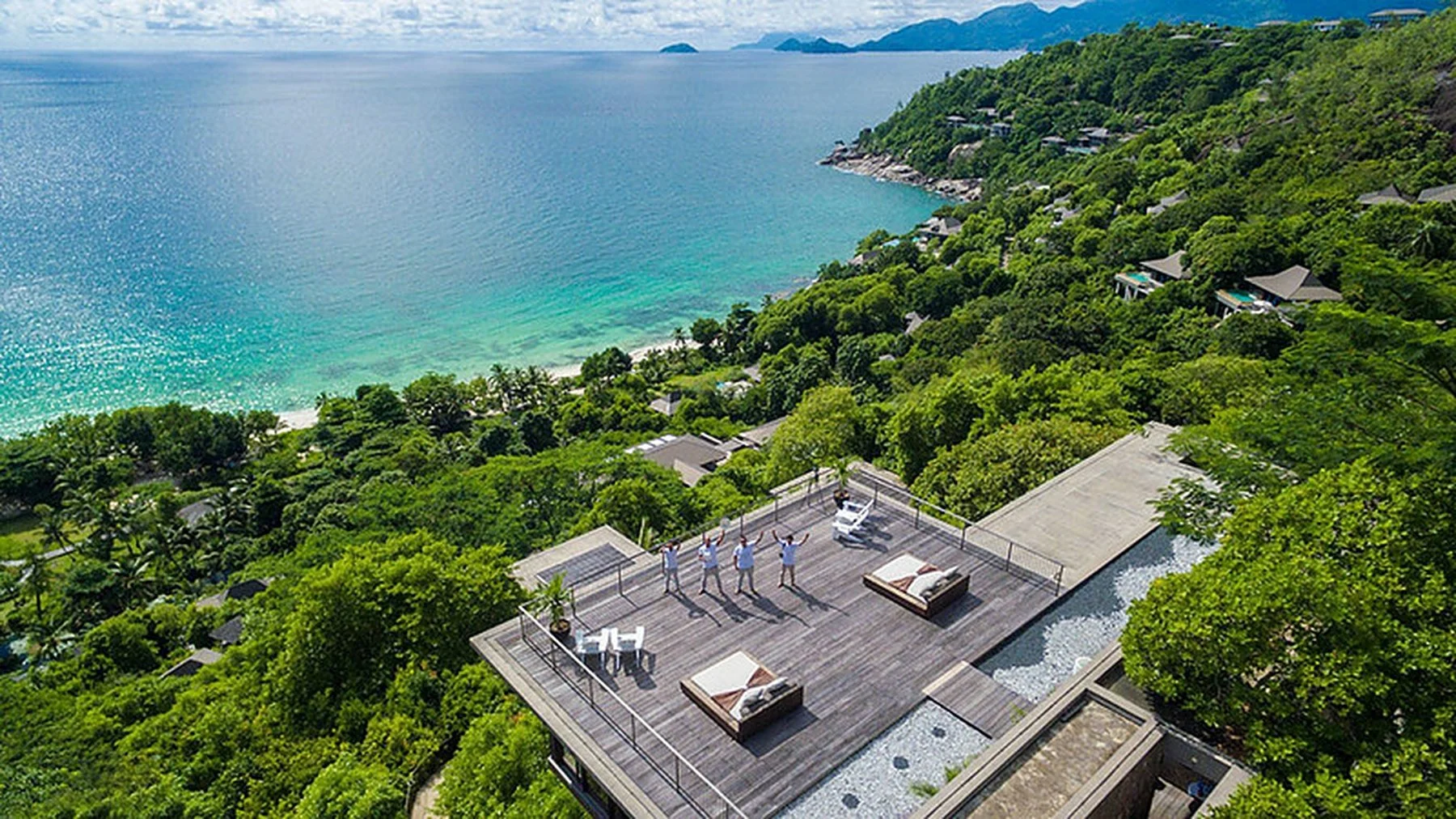 A rooftop deck overlooking a tropical coastline with lush green hills, ocean, and distant islands, featuring five people celebrating with raised hands, lounge chairs, and yoga mats.