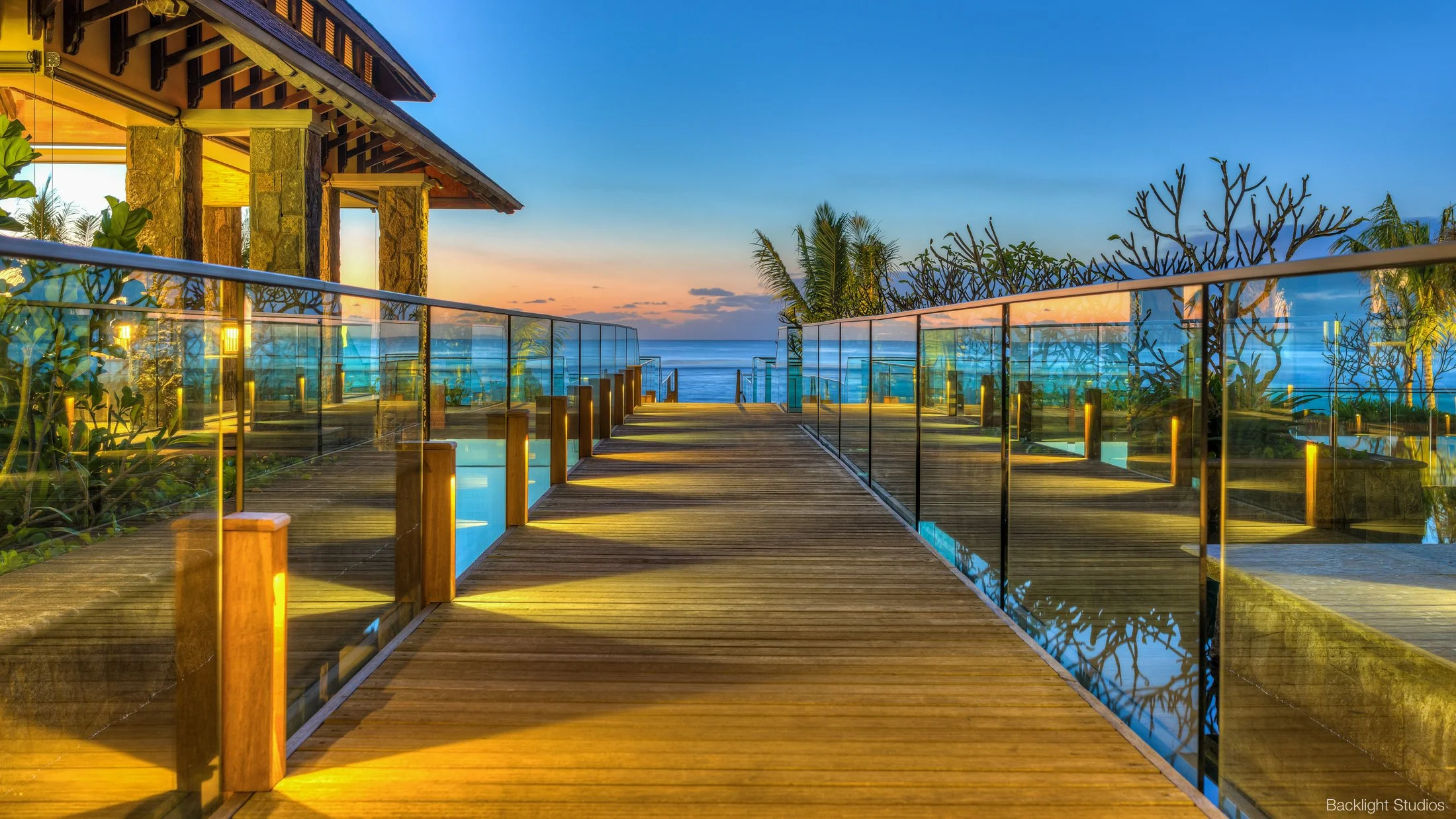 A wooden walkway with glass railings leads toward the ocean under a colorful sunset sky with orange and blue hues. There are tropical plants and trees on either side of the walkway, and a building with stone columns and a sloped roof on the left. The scene is illuminated with warm lighting from the building and pathway.