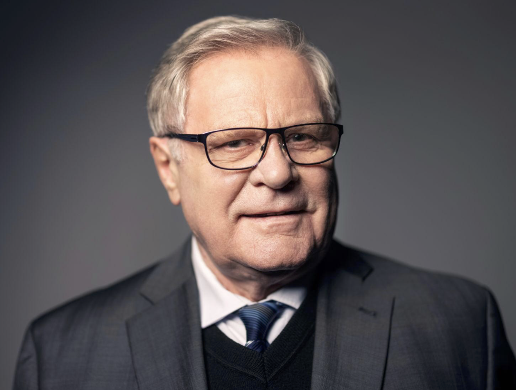 Headshot of a middle-aged man with glasses, gray hair, wearing a dark suit and tie, against a dark gray background.