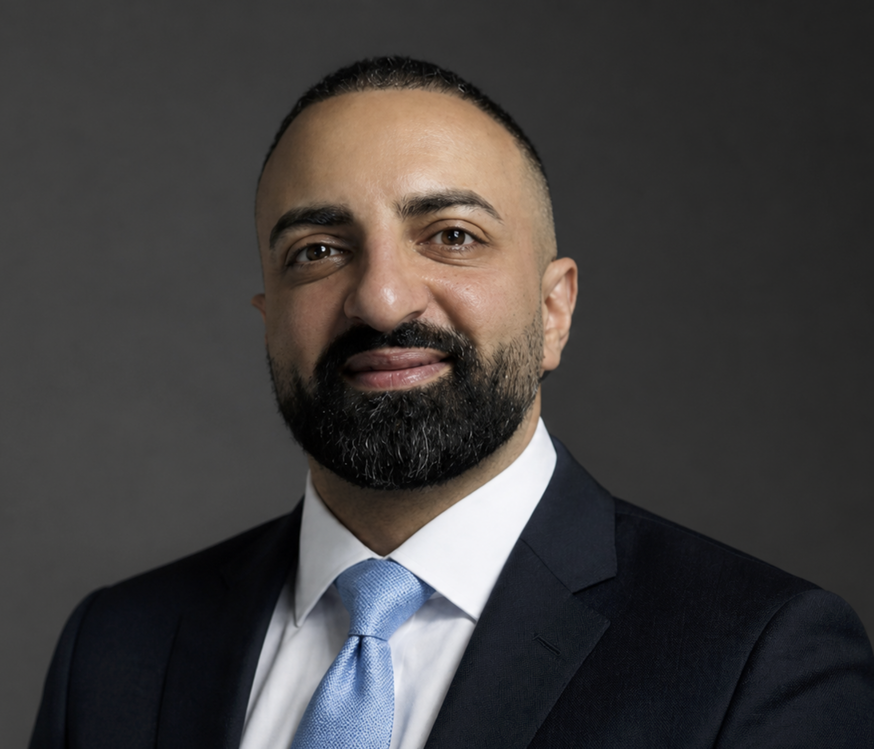 A man with a dark beard and mustache wearing a dark suit, white dress shirt, and light blue tie, with a neutral gray background.