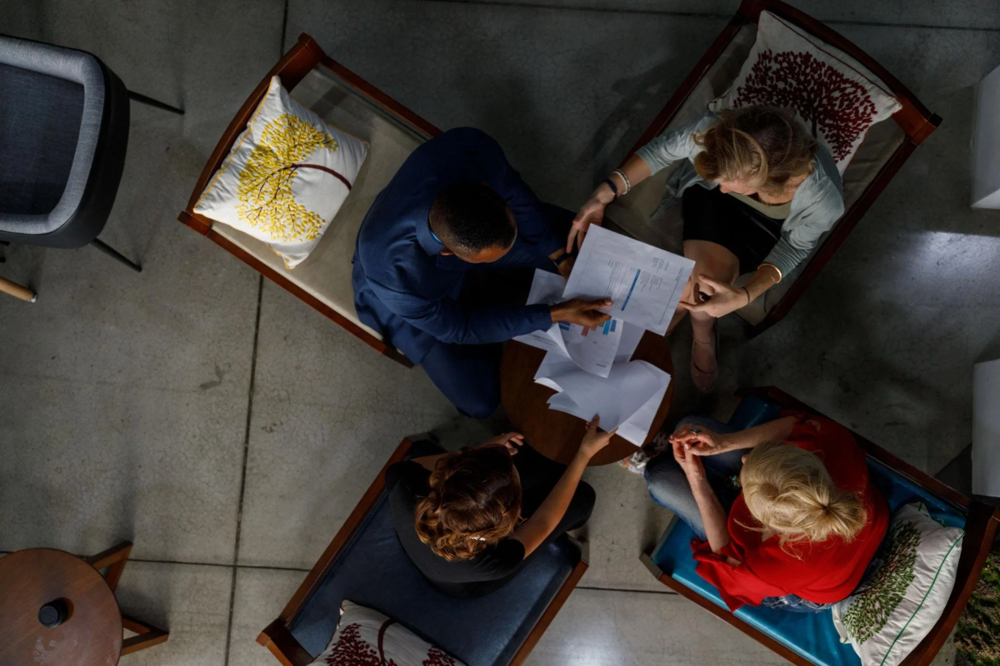 A top-down view of a meeting with several people sitting around a wooden table, looking at documents. Two empty chairs with embroidered pillows are at the sides of the table.