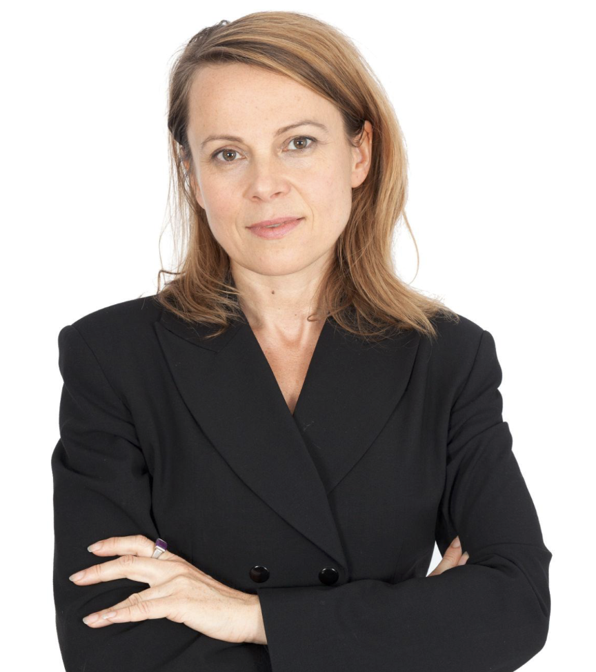 A woman with shoulder-length reddish-brown hair, wearing a black blazer, standing with arms crossed against a white background.