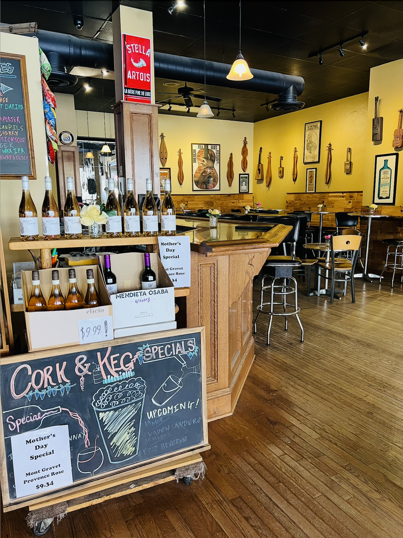 Interior of a restaurant or bar with musical instrument wall decorations, tables with small flowers, and a chalkboard sign advertising cork and keg specials with wine bottles on display.