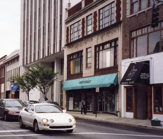 Street view with parked cars, a storefront with a teal awning, and buildings with large windows and trees in the background.