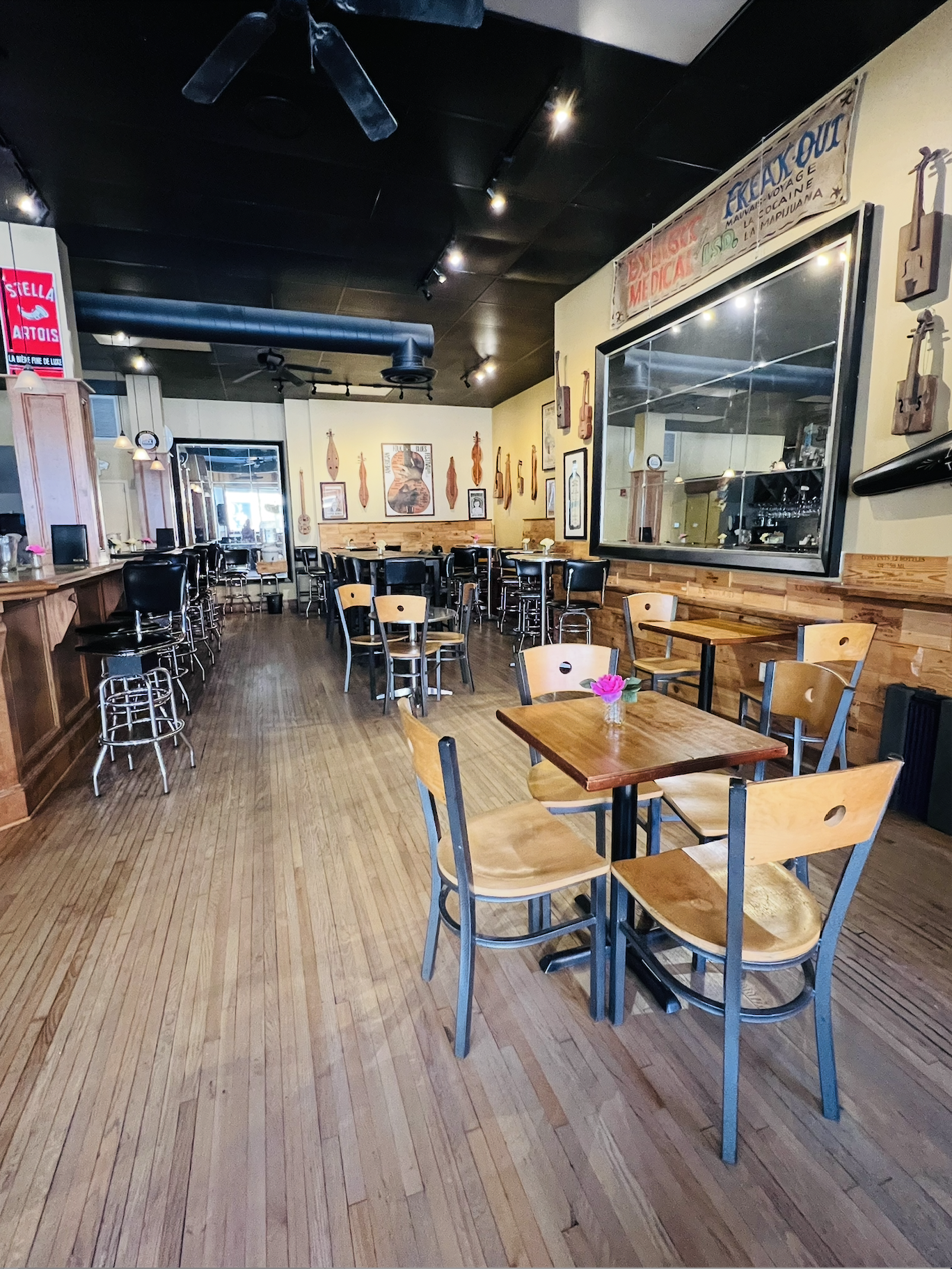Inside of a cozy restaurant or café with wooden floors, tables, and chairs. The walls are decorated with musical instruments and framed pictures. There is a large window or mirror on the side. The ceiling is painted black with track lighting and a ce